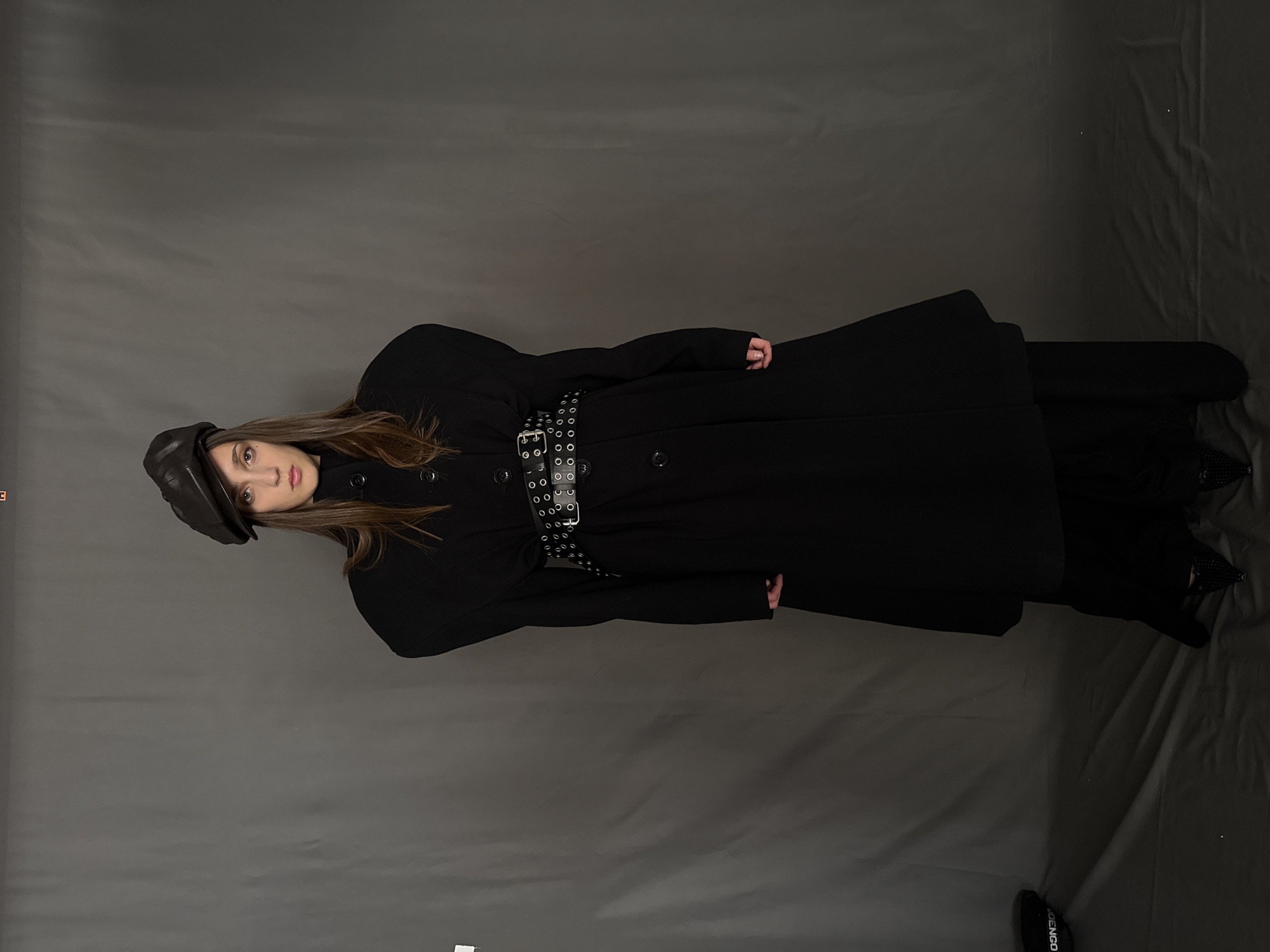 A young woman poses in a studio against a gray backdrop. She wears a black newsboy cap, a long black coat with visible buttons, and a black belt with circular metallic embellishments around her waist. The coat appears to be cinched by the belt. She also seems to be wearing black pants and shoes.