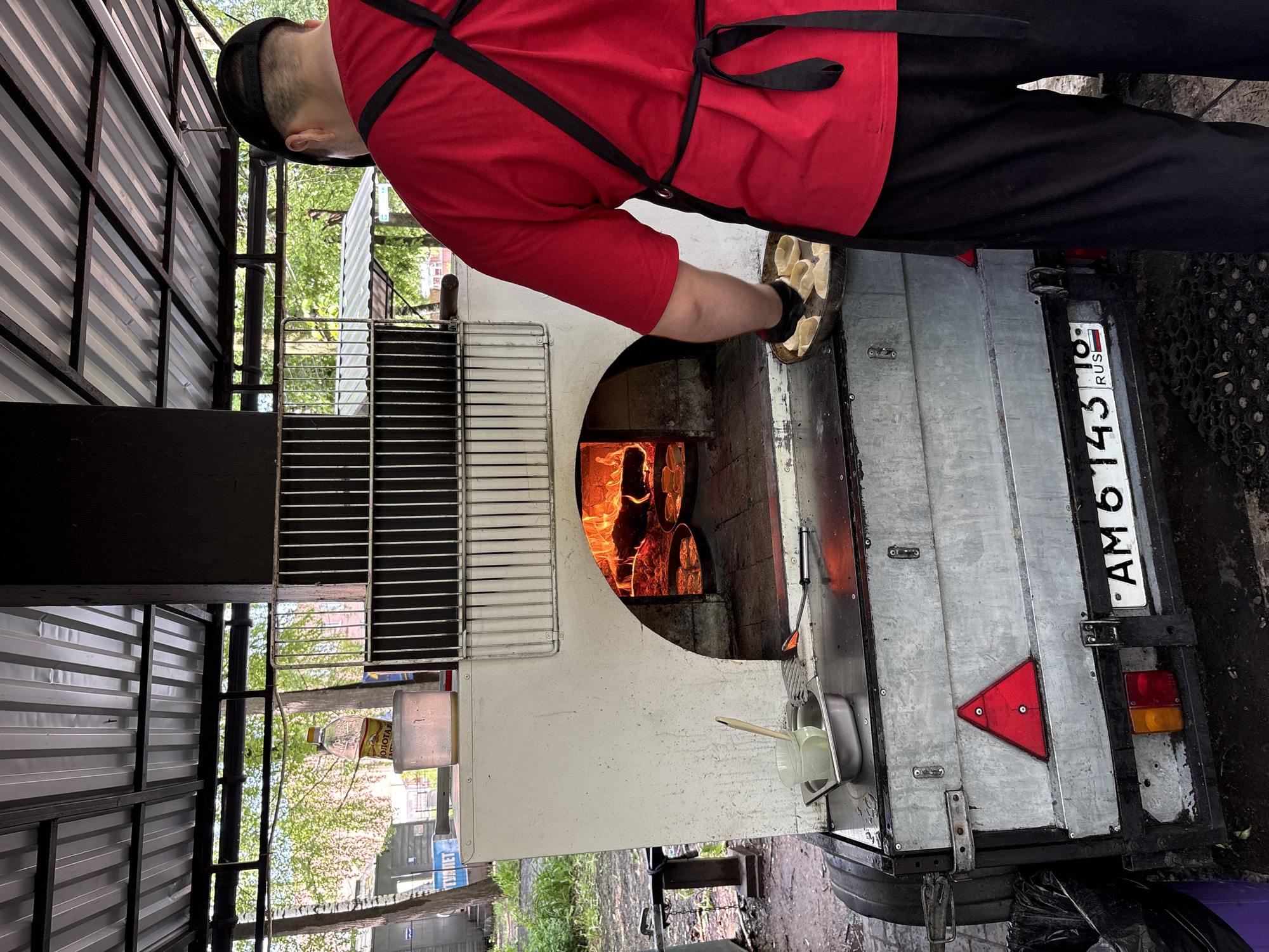 A person in a red shirt and black apron is putting a tray of food into a wood-fired oven mounted on a trailer. The trailer has a Russian license plate that reads "AM 6143 18 RUS." The trailer is parked in what appears to be an outdoor market. There is a metal grate to the left of the oven and a container with utensils hanging from the trailer. Based on the license plate region code "18," this image was likely taken in Izhevsk, Russia.
