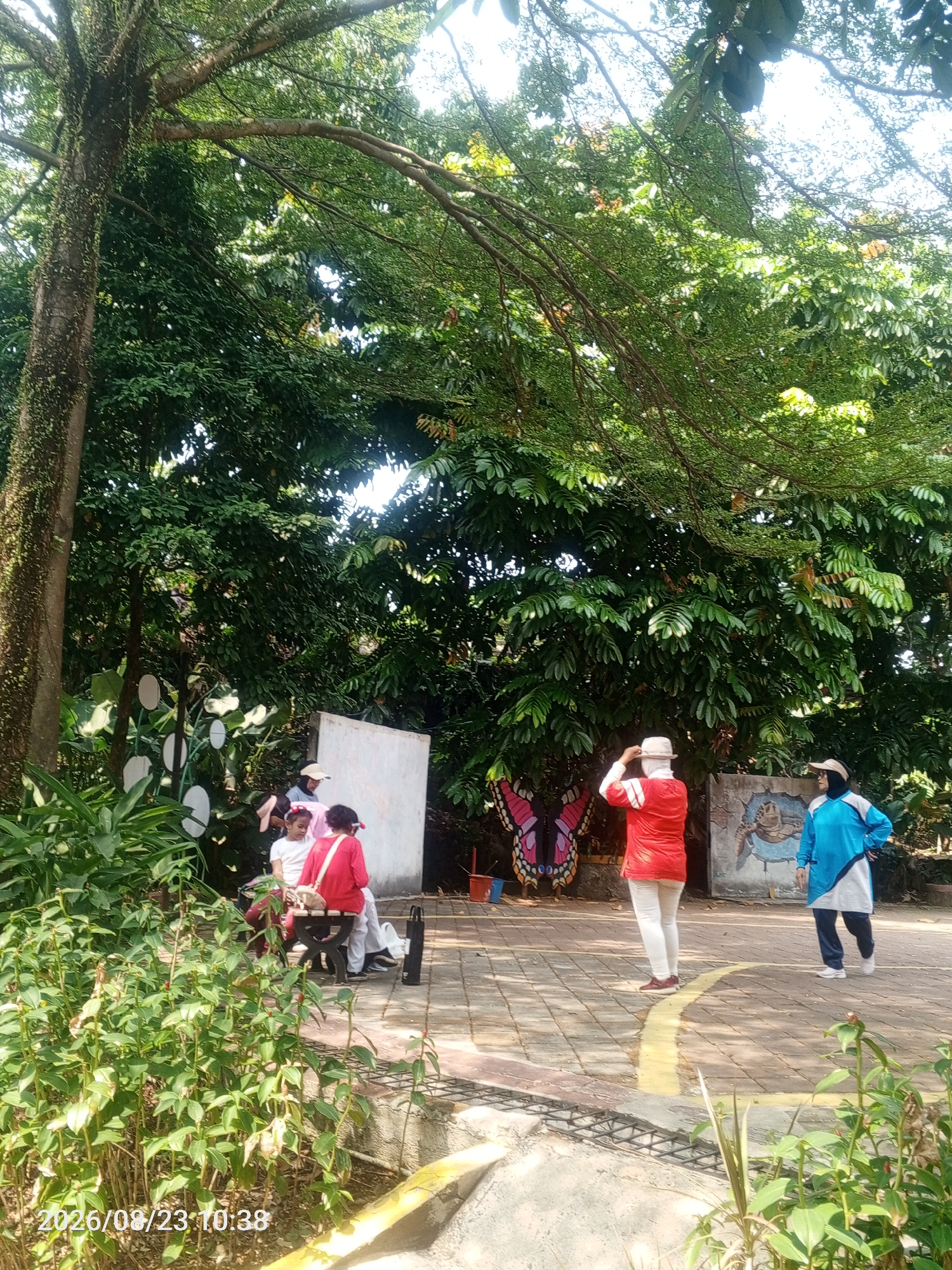 In Depok, Indonesia, a group of individuals are gathered outdoors in what appears to be a park or recreational area. The scene is dominated by lush green foliage and mature trees, with sunlight filtering through the canopy. In the foreground, several people are present, including two young girls in pink and red attire, one of whom is seated. To their left, a white wall is visible, featuring a large pink and black butterfly decal.
Further back, two adults are interacting. One individual, wearing a red top and white pants, is facing away from the camera, adjusting a light-colored hat. Beside them, another person in a blue and white athletic outfit stands with their hands on their hips, looking towards the left. Behind this individual, a mural depicting a turtle within a cracked wall can be seen. The ground is paved with interlocking bricks, marked by a yellow line. The overall ambiance suggests a casual outdoor activity or gathering.