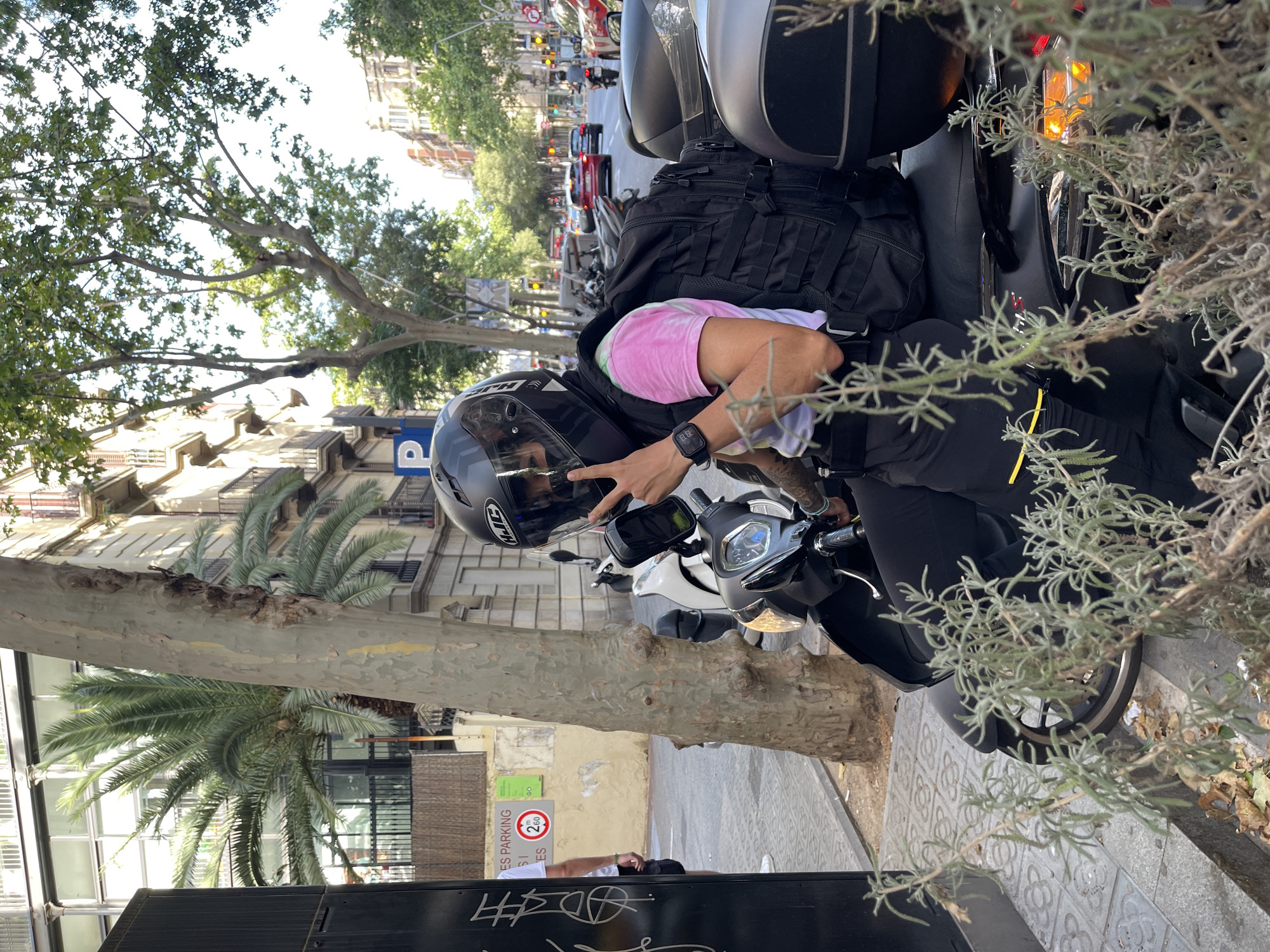 A person wearing a black helmet with "HJC" branding and a pink and white tie-dye t-shirt is sitting on a black scooter. The person is holding up two fingers in a peace sign with their right hand. They are wearing a black watch on their left wrist and have a large black backpack strapped to their back. The scooter has a black seat and what appears to be a grey storage compartment on the back. In the background, there is a tree with a textured trunk, a building with balconies and windows, and other scooters and cars on a street. A street sign with a "P" symbol and a circle with "2.60" inside is visible on the left side of the image. The pavement is tiled, and there are plants in the foreground. The scene appears to be set in Barcelona, Spain, as indicated by the architecture and street elements typical of the city.