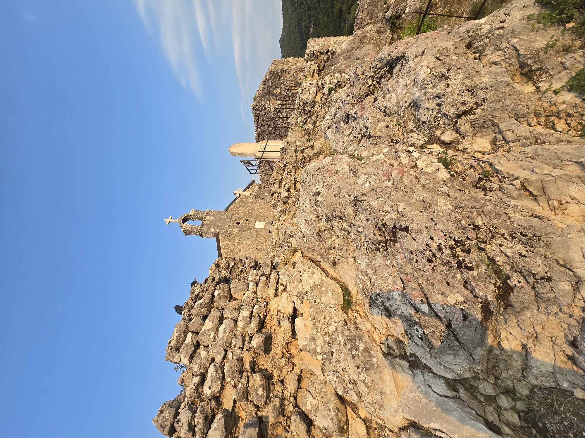The image shows the Church of St. George, located on a rocky outcrop overlooking the village of Gornja Vrbica, Montenegro. The church, built of stone, has a bell tower topped with a cross. In the foreground, rough, weathered rocks dominate the scene, with patches of green vegetation growing in crevices. The sky is a clear blue, with a few wispy clouds. The overall impression is one of rugged beauty and isolation.