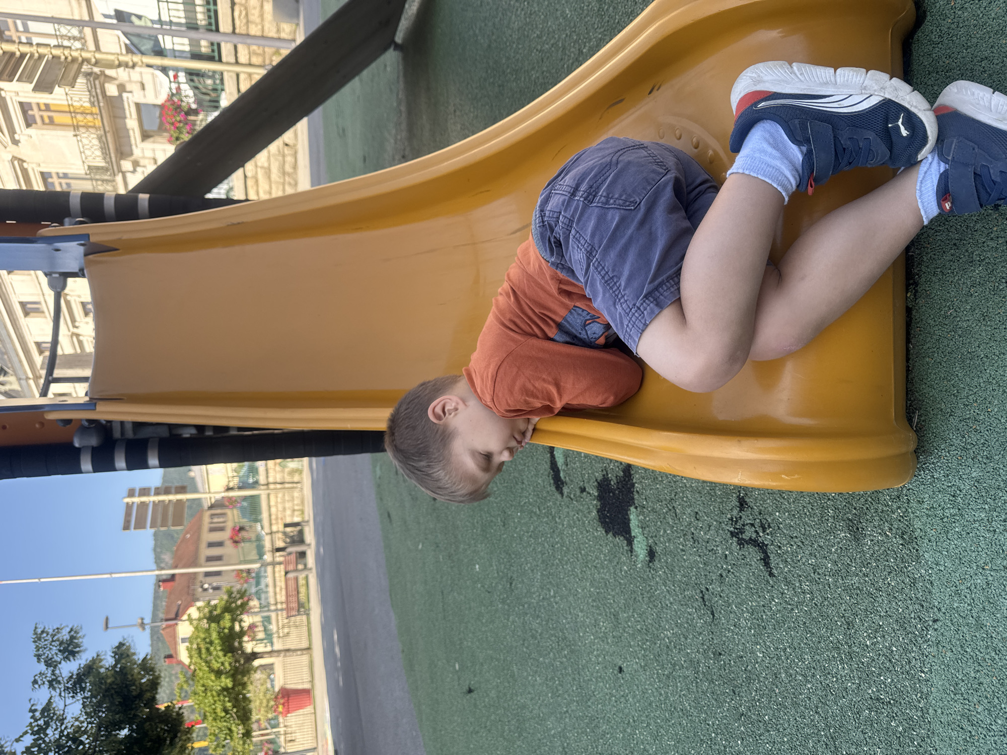 A young boy is sleeping on a yellow slide at a playground in Saint-Claude, France. He is wearing an orange long-sleeved shirt, blue shorts, and blue and white sneakers. The playground has a green rubberized surface. In the background, there are trees, buildings, and a clear blue sky.