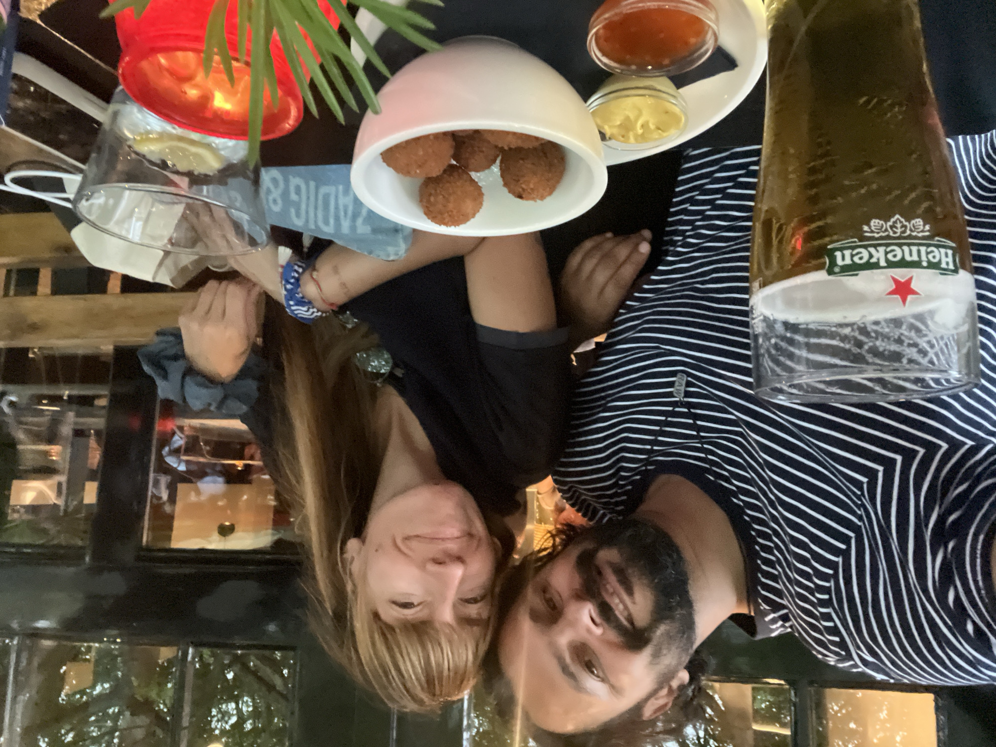 This is a photograph of two people, a man and a woman, smiling at the camera. They are seated at a table, and the image is framed from above, as if looking down on them. The scene appears to be outdoors, possibly on a patio or terrace, with trees and reflections visible in the background. The lighting suggests it is daytime, though perhaps late afternoon or evening due to the warmth of the light.
On the table, there are several items. A red bucket with what appears to be a drink or ice is visible, along with a glass containing a pale liquid, possibly water with a slice of lemon. A white bowl holds a serving of fried, ball-shaped snacks, likely bitterballen, a Dutch delicacy. Beside the bowl are two small dishes containing dips, one yellow and one reddish. A bottle of Heineken beer, partially filled, sits in the foreground.
The couple is dressed casually. The woman has long, blonde hair and is wearing a dark top. The man has dark hair and a beard and is wearing a horizontally striped shirt. Both appear to be relaxed and happy, sharing a moment together over food and drinks.
A partial sign with the text "ZADIG & VOLTAIRE" is visible, which is a French fashion brand. Given the presence of bitterballen and Heineken, and the location context provided as Delft, Netherlands, the scene likely takes place at an outdoor cafe or restaurant in Delft.
