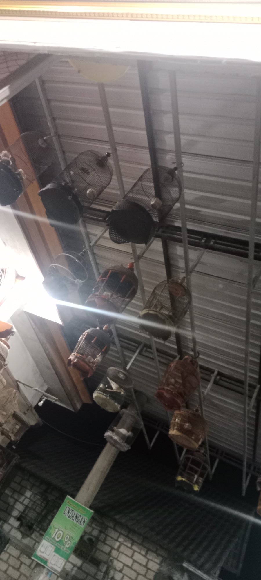 In Rengel, Indonesia, a low-angle, slightly tilted shot captures a collection of birdcages hanging from a metal roof structure. The birdcages are of various sizes and materials, with some appearing to be made of dark metal and others of lighter wood. A few of the cages have black coverings at the bottom. In the lower left of the frame, a green sign with Indonesian text, including "UNDANGAN" and "10%", is visible, attached to a light-colored pole. The background consists of a corrugated metal roof and brickwork. The lighting is somewhat dim with some bright highlights, suggesting it may be indoors or under a covered area. No people are visible in the image.