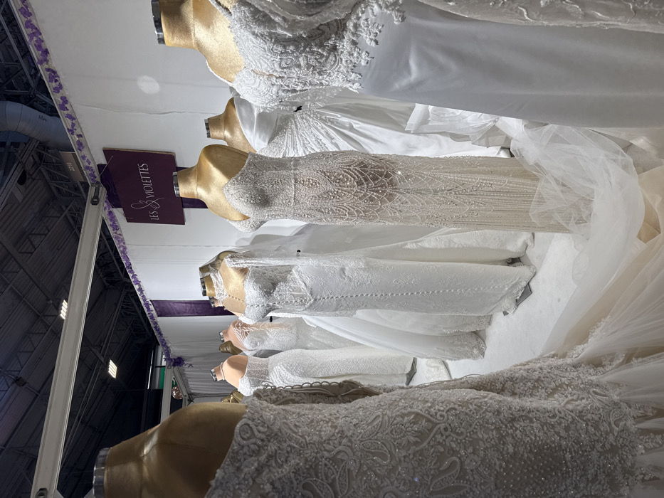 This image depicts an indoor display of approximately ten headless mannequins exhibiting various white and off-white wedding dresses. The scene is set within an exhibition hall or trade show, identifiable by the dark industrial ceiling with exposed ducts, girders, and fluorescent lighting. The specific location is a vendor booth, likely in Paris, France, given the French signage and the request in the prompt.
The mannequins feature gold-colored upper torsos and stand on a light-colored platform or floor. The dresses display diverse styles, including form-fitting silhouettes, A-line cuts, and gowns with extensive trains made of tulle and lace. Intricate details such as elaborate beading, sequins, embroidery, and delicate lace patterns are prominent on most garments. One dress on the far left shows a complex paisley-like pattern. Another in the center features a strapless sweetheart neckline with dense vertical beading, while a neighboring dress has a high, beaded collar and long, sheer, embellished sleeves.
The booth's background consists of white partition walls, adorned along the top edge with purple fabric strips and floral-like purple garlands. A prominent purple sign with white cursive text reads "LES VIOLETTES." A smaller number, "221D," is visible on the upper white wall. No people are present in the frame; the scene is static, focused entirely on the apparel and display setup.