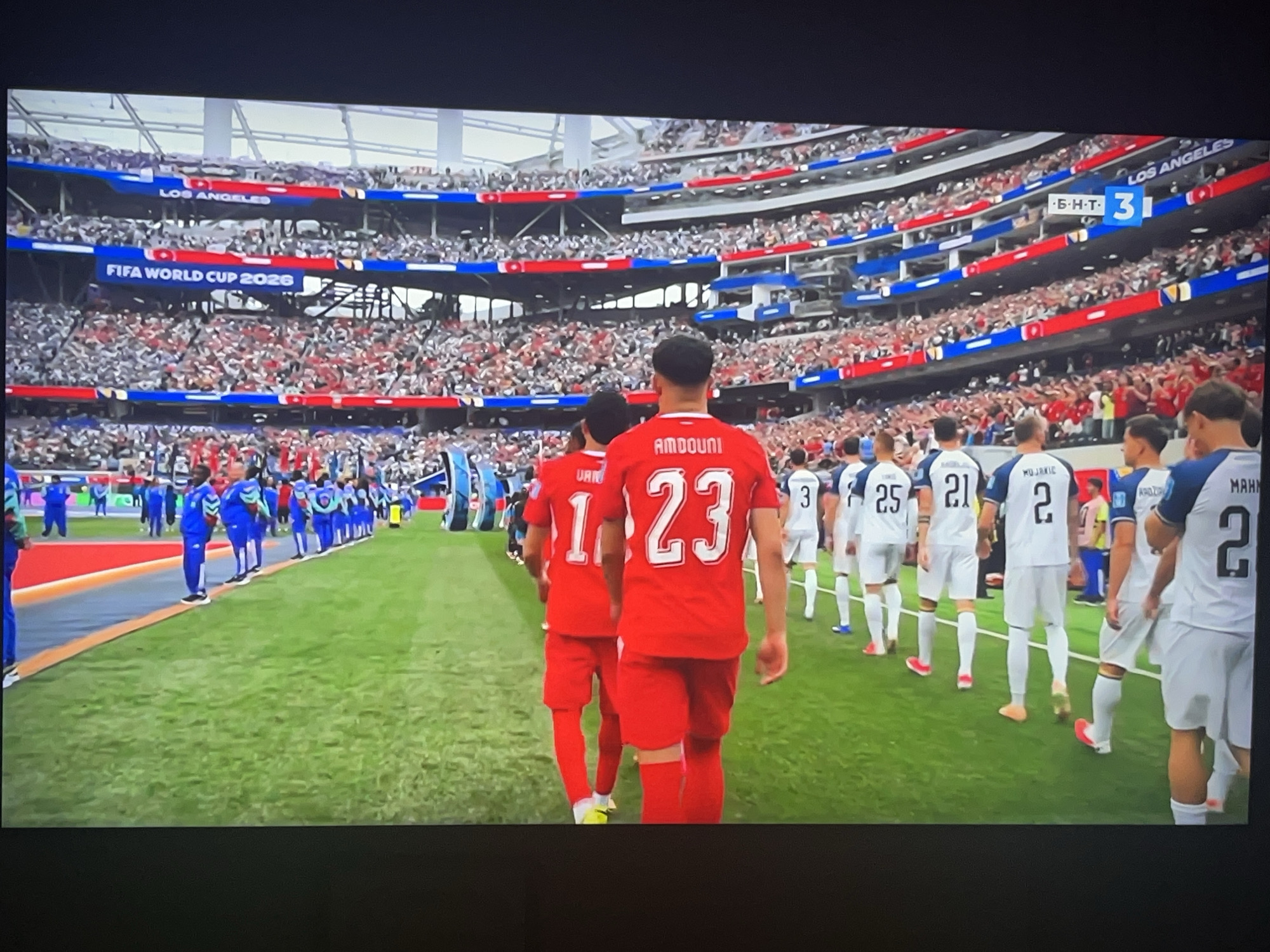 A high-angle, wide shot captures a soccer match in progress at a stadium, with the score display indicating a 3-0 lead for one team. The stadium seating is packed with spectators, predominantly in red and white, arranged in sections separated by blue banners. A prominent banner in the stands reads "FIFA WORLD CUP 2026 LOS ANGELES". On the vibrant green field, players from two teams are visible. One team, in striking red jerseys with white numbers and trim, walks in a procession. The other team, dressed in white jerseys with blue trim and numbers, stands in a line. The players in red have names on the back of their jerseys; one reads "AMDOUNI" above the number "23". The players in white have names like "MUJAKIĆ" and numbers such as "2" and "21". The overall scene is dynamic and indicative of a significant sporting event. The mention of Varna, Bulgaria, in relation to this image is incongruous with the visible stadium signage and branding clearly indicating Los Angeles and FIFA World Cup 2026.