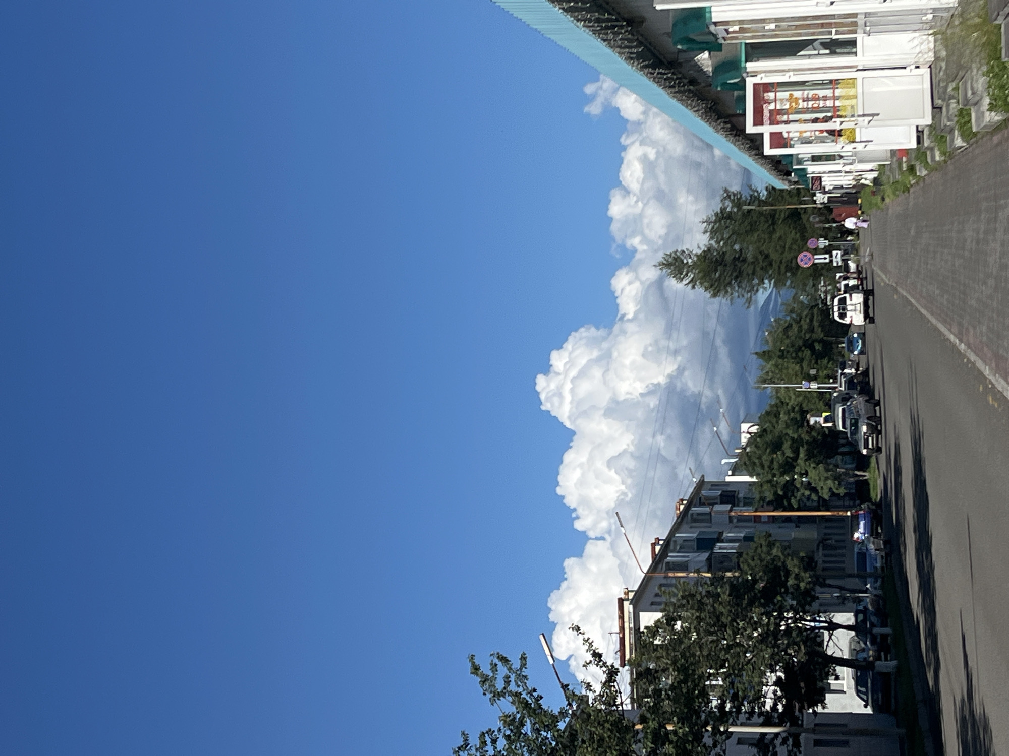 The image captures a street scene in Petropavlovsk-Kamchatsky, Russia, under a bright blue sky with scattered cumulus clouds. On the left side of the street, a multi-story apartment building is visible, lined with trees. Several cars are parked along the street and in front of the building. The right side of the street features a row of shops with white facades and brightly colored entrances, many with awnings. A sidewalk runs along the shops, and a few pedestrians are walking on it. Trees and a traffic light are also present in the scene. The overall impression is of a typical urban environment on a clear day.