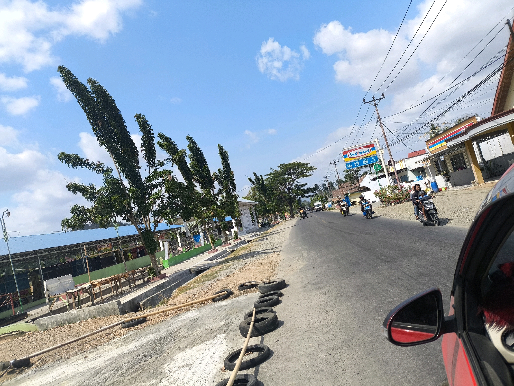 The image depicts a street scene in Talumopatu, Indonesia, under a bright blue sky with scattered white clouds. Tall, slender trees line the left side of the road, some with a distinctive upright growth pattern. Adjacent to the trees is a building with a blue roof and outdoor seating areas with wooden tables and benches, suggesting a restaurant or market.
The road itself is asphalt and appears to be a two-lane street with some traffic. Several motorcycles are visible on the road, with riders. One motorcycle is in the foreground on the right, its rider wearing a dark jacket, and another behind it with a passenger. Further down the road, more motorcycles are seen.
On the right side of the road, there are buildings with shops. A prominent sign for "Indomaret," a convenience store chain, is visible, along with utility poles and a complex network of electrical wires.
The foreground on the left shows a dirt and gravel area with several old tires placed along the edge of the road, some with bamboo poles threaded through them, possibly serving as a barrier or for some other roadside purpose. The right side of the image shows the side mirror and part of a red car, indicating the photograph is taken from inside a vehicle. The overall impression is that of a typical roadside scene in a populated area of Indonesia on a sunny day.