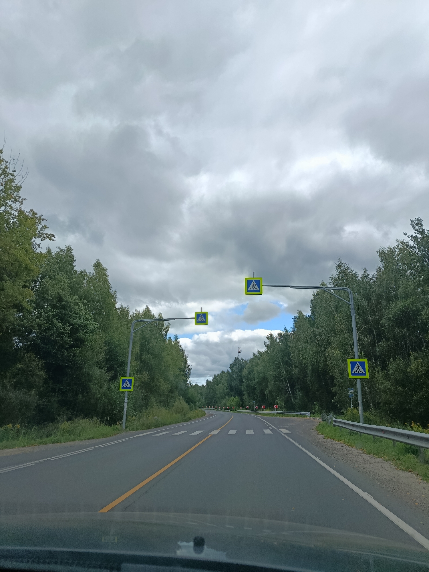 The image depicts a view from a vehicle driving on a paved road in a rural area, likely on the outskirts of 1-Go Maya, Russia. The road is lined with dense green trees on both sides, suggesting a forested environment. The sky is overcast with heavy grey clouds, but there are some patches of blue visible.
The road has two lanes, marked with a solid yellow line down the center and white lines separating the lanes. There are multiple pedestrian crossing signs, indicated by blue square signs with a white triangle featuring a pedestrian symbol. Some of these signs are mounted on poles along the side of the road, while others are suspended over the road on metal gantries.
There are also road signs indicating sharp turns ahead, with red and white directional arrows visible in the distance on the right side of the road. The presence of these signs and the clear markings on the road suggest it is a well-maintained public road.
The foreground of the image shows the dashboard of the vehicle from which the photo is taken, indicating the perspective of a driver or passenger. No people are visible in the scene. The overall atmosphere is one of a cloudy, late summer or early autumn day in a rural landscape.
