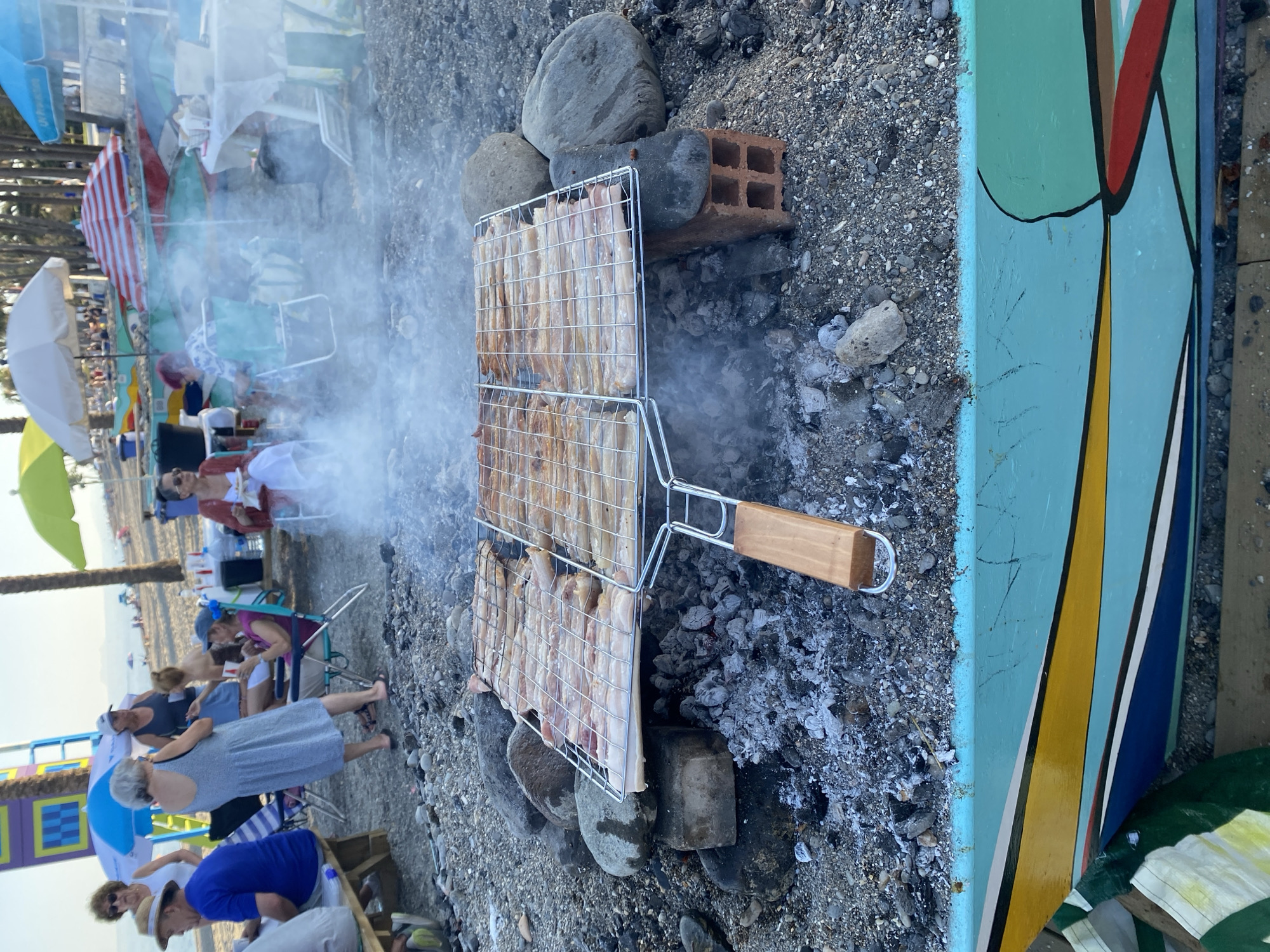 The image captures a lively scene on a beach in Marbella, Spain, possibly during a casual gathering. In the foreground, a barbecue grill with several rows of meat is actively cooking over hot coals, generating a visible plume of smoke. The grill is precariously balanced on rocks and a brick. Behind the grill, a group of people are enjoying themselves in a relaxed atmosphere. Some are seated in beach chairs, while others are standing and chatting. Umbrellas, some brightly colored, dot the sandy beach, providing shade. In the background, the calm sea and a distant coastline suggest a typical Mediterranean setting. The overall impression is one of a pleasant outdoor meal or celebration on a sunny day.
