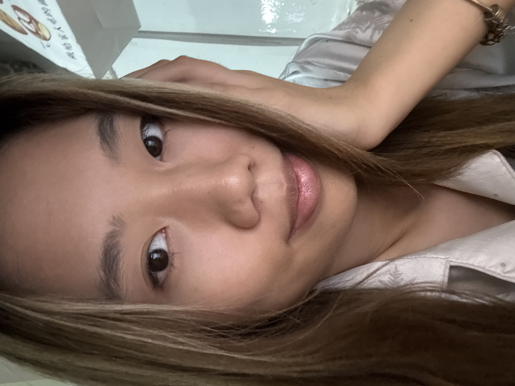 A close-up, selfie-style portrait shows a woman with light brown hair and brown eyes. She is wearing a light-colored shirt with a small design near the collar. A bracelet is visible on her arm. The background is indistinct, with a white surface and blurry text. The photograph's location in the Nan'an District of China is unknown.