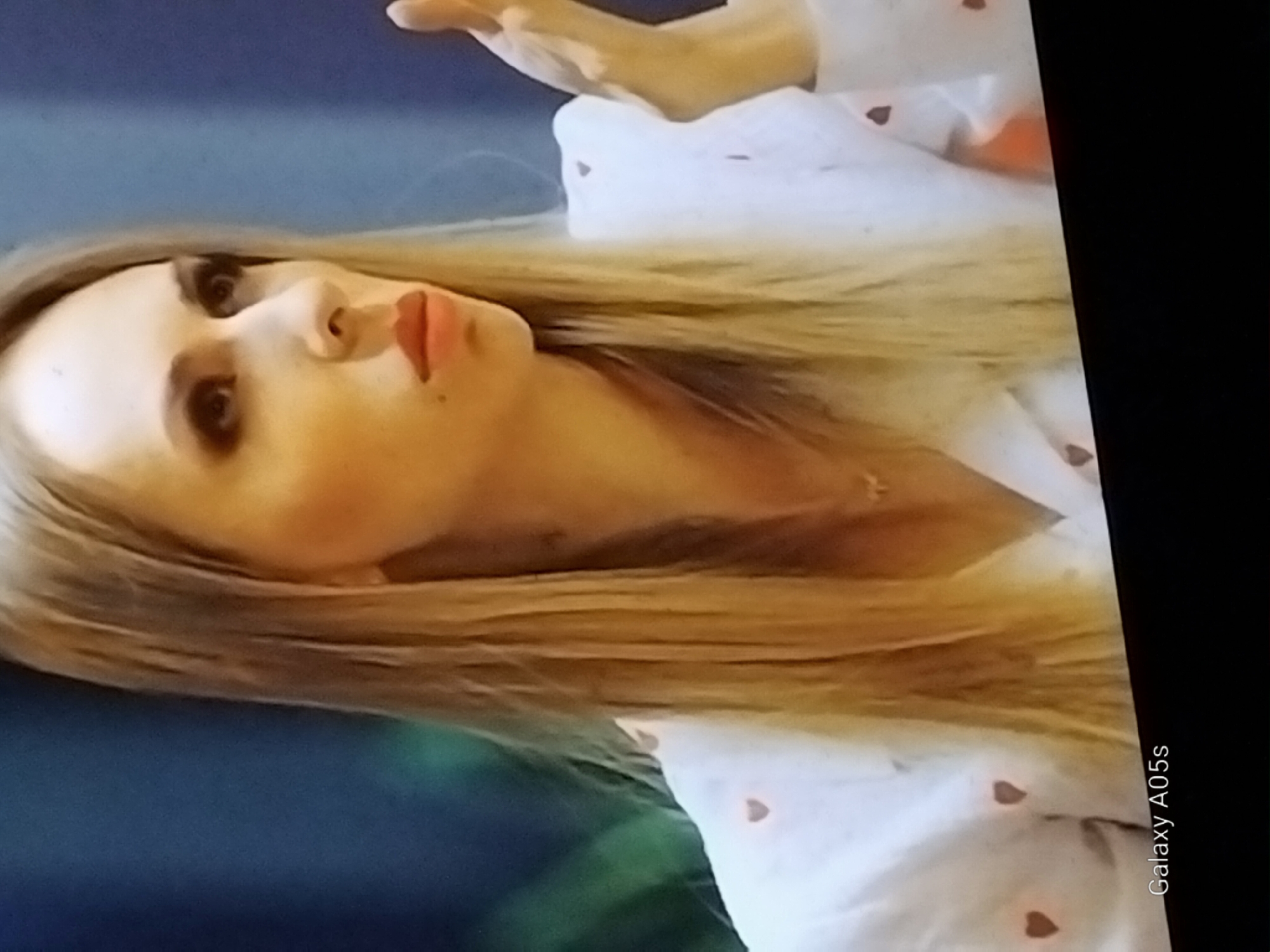 The image shows a medium close-up of a woman with long, blonde hair. She is wearing a white top with a subtle heart pattern. Her makeup includes dark eyeshadow and lipstick. Her facial expression suggests she is speaking or reacting to something. Her left hand is raised, with fingers spread. The background is blurred and appears to be indoors with some greenery visible. Text at the bottom of the image reads "Galaxy A05s". The location in the city of Druzhba, Russia, cannot be determined from the visual information provided in the image.