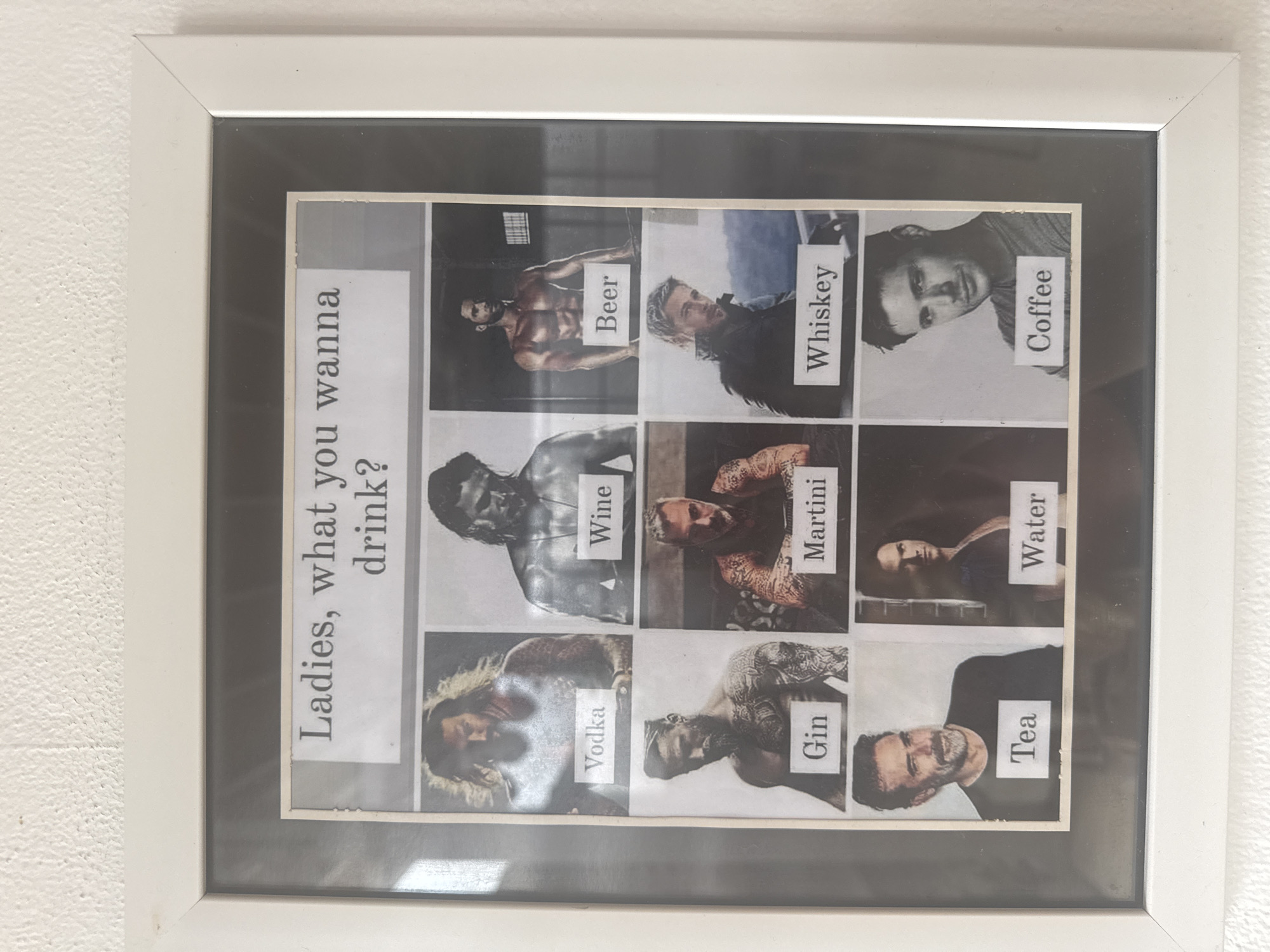 A framed picture hangs on a white wall. The picture is a collage of black and white photographs of muscular men, each paired with a different beverage name: "Beer", "Whiskey", "Coffee", "Wine", "Martini", "Water", "Vodka", "Gin", and "Tea." The text "Ladies, what you wanna drink?" is printed on the collage as well. The frame is white, with a gray border. There is no indication of Nusa Penida, Indonesia.