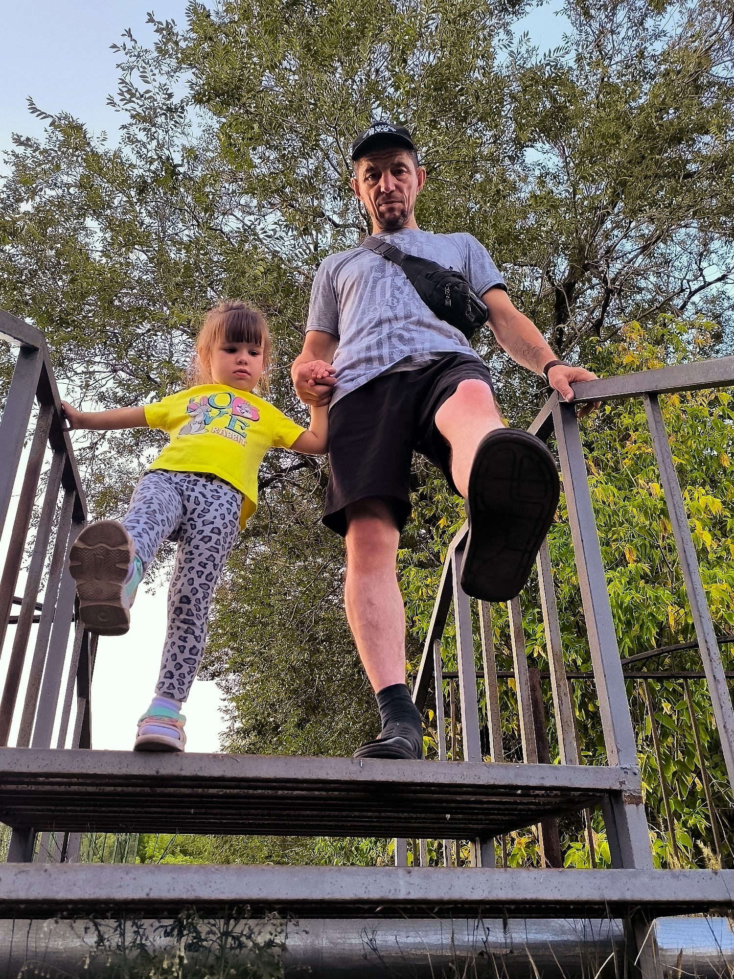 A low-angle shot captures a man and a young girl standing on a metal outdoor staircase, looking upwards towards a canopy of green trees. The man, wearing a grey t-shirt, black shorts, a black baseball cap, and a black fanny pack across his chest, has his right leg raised, stepping upwards on the staircase. He is holding the girl's left hand with his right hand. The girl, on the left side of the frame, wears a yellow t-shirt and grey leopard-print leggings. Her left leg is also raised, stepping onto a higher stair. Her right hand is resting on the metal railing. The background consists of lush green foliage and a clear sky. The overall scene appears to be in an outdoor, possibly urban park or recreational area within the city administration of Qarağandı, Kazakhstan.