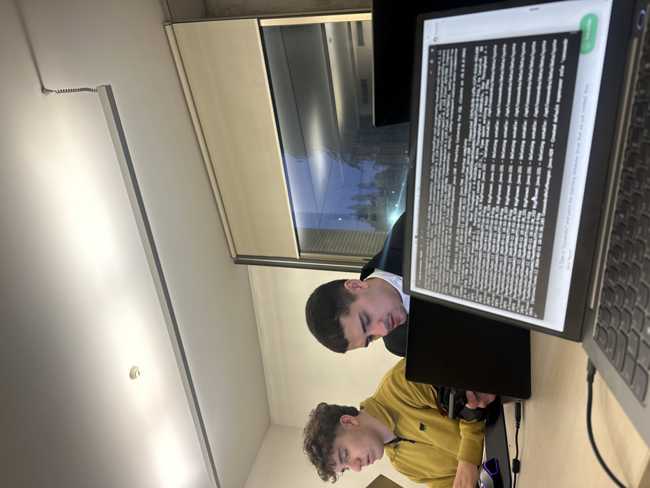The scene depicts an interior workspace within the city of Guimarães, Portugal. Two young adult males are seated at light-colored desks, engaged with individual laptop computers.
The individual positioned on the left has short, curly dark brown hair and is wearing a mustard yellow long-sleeve polo shirt with a small dark emblem on the left chest. His gaze is directed downwards, and his right hand rests on a black computer mouse situated on a dark mousepad that exhibits a subtle purple LED glow.
The individual in the center has short, dark hair and is wearing a dark jacket over a white collared shirt. He is looking down at a black laptop screen directly in front of him.
In the foreground, a silver-colored laptop is visible, its screen displaying an active terminal interface with technical text. The content includes references to "Cassandra" and "Java," suggesting programming or system administration activities. A black cable is connected to the left side of this laptop.
The environment includes a white ceiling with recessed linear lighting fixtures and a white circular smoke detector. A coiled wire descends from the ceiling near the windows. Large windows line the background, partially covered by beige roller blinds. Reflections of the interior lighting are visible on the glass, behind which a dark, indistinct outdoor scene featuring trees and distant lights is discernible, indicating evening or nighttime conditions. The walls are light-colored.