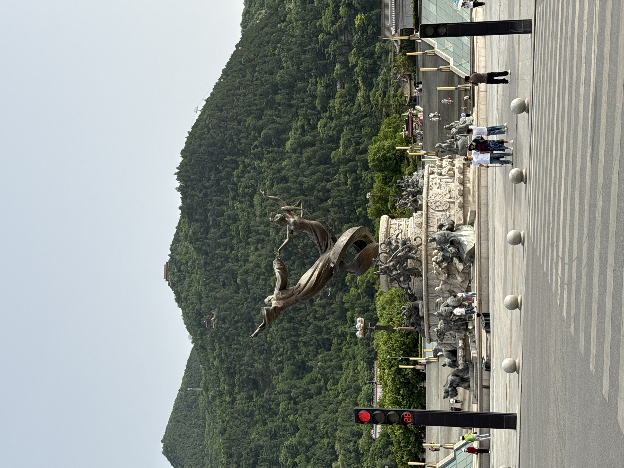 A vertical, medium shot shows a large monument in Lintong District, China, surrounded by a paved plaza, with a mountain in the background.
The monument features bronze-colored statues of people in dynamic poses, some appearing to be embracing or reaching for each other. The monument rests on a wide, decorated platform with additional sculpted figures. People are walking along the plaza near the monument. Some are walking across a crosswalk.
To the right of the monument, there is a traffic light showing a red light and a digital countdown timer displaying "28".
In the background, a mountain covered in lush green trees rises. There is a structure visible on the mountainside, and a cable car lift. The sky above the mountain is overcast and gray.