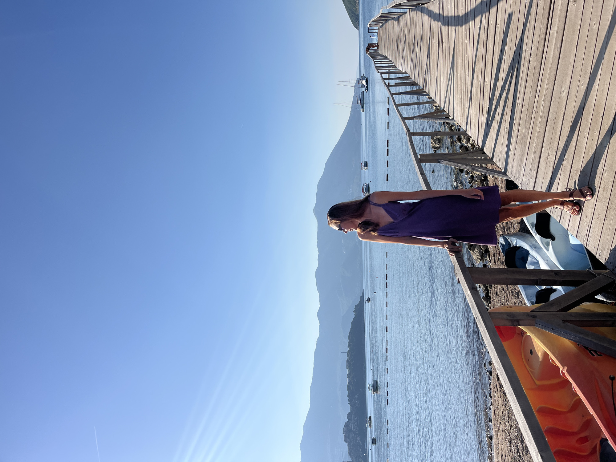 A young woman stands on a wooden pier extending into a calm, blue sea under a clear sky. She is wearing a purple sundress and brown sandals, looking to her left with sunglasses on. The pier has a wooden railing and casts long shadows. In the background, several boats are anchored on the water, and mountains rise in the distance. In the foreground, near the shore, two kayaks, one orange and one blue, are visible. The scene appears to be a coastal area, possibly near Budva, Montenegro, given the mountainous coastline and clear blue water often associated with the Adriatic Sea. The sun is shining brightly, suggesting it is a warm, sunny day.
