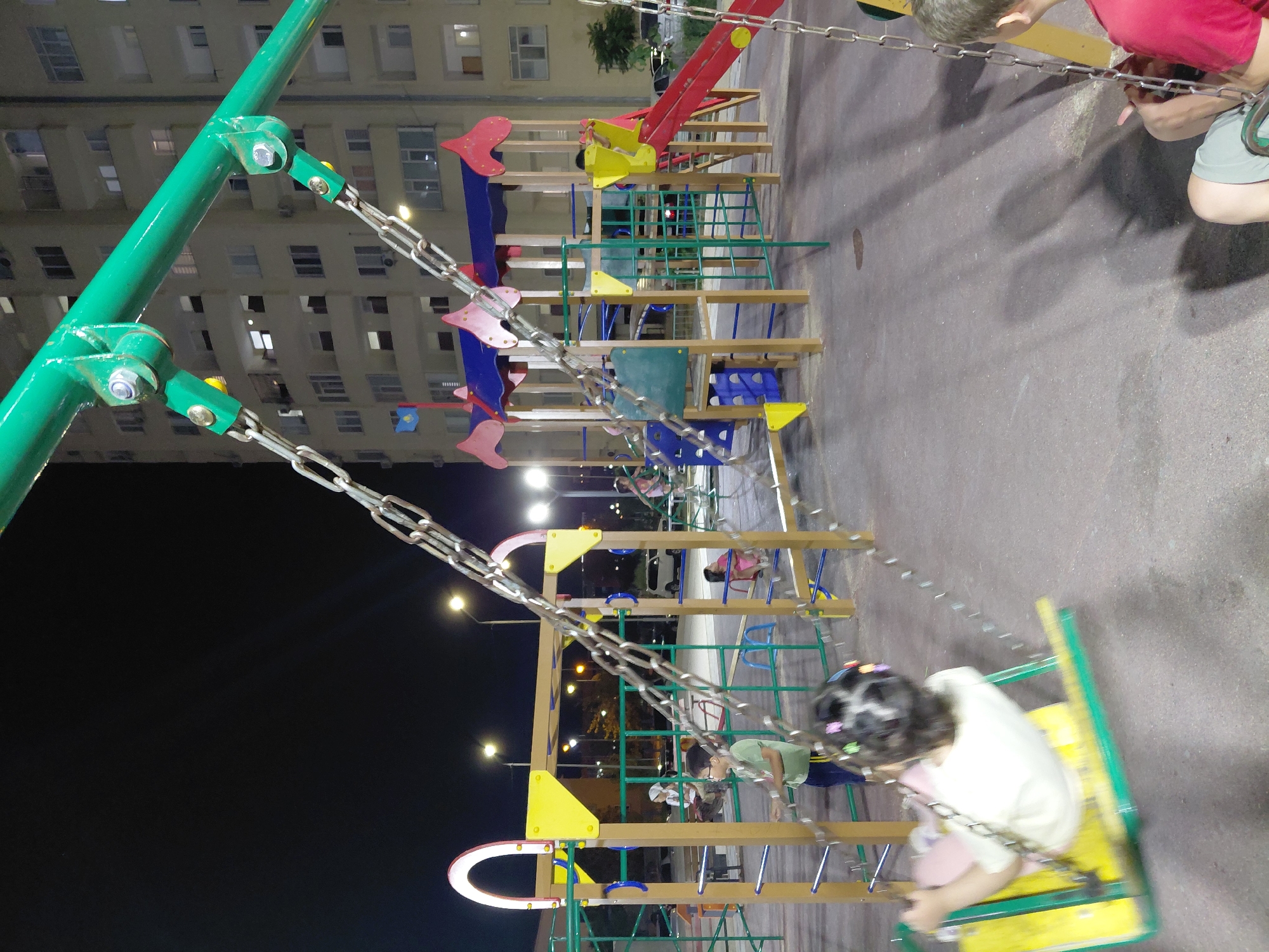 In Aktau, Kazakhstan, a nighttime scene unfolds at a children's playground in front of a multi-story residential building. The playground features a green metal swing set with a girl on a yellow and green swing in motion, her hair adorned with colorful clips. Nearby, a boy in a green shirt and dark pants stands by the swing set. Another child in a pink dress is visible on a play structure in the background. The playground equipment includes slides, climbing nets, and colorful wooden structures. Streetlights illuminate the area, casting long shadows on the asphalt ground. The residential building behind the playground has lit windows, indicating occupancy. The overall scene depicts a typical evening at a community playground.