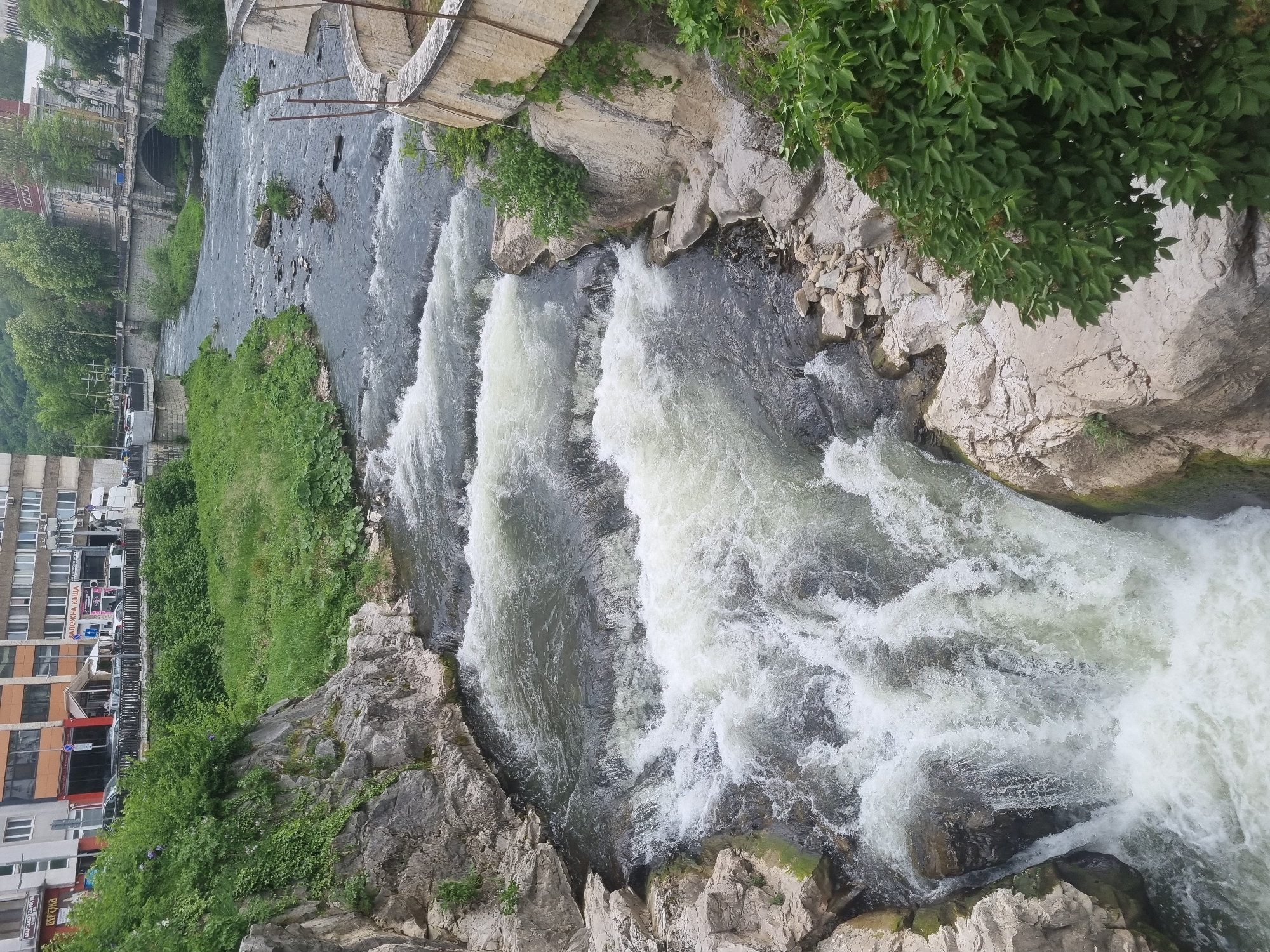 The image shows a cascading river in the city of Gabrovo, Bulgaria. The water flows over several stepped sections, creating white foamy water in contrast to the darker water above. On the left side, green vegetation grows along the riverbank with an adjacent building visible. On the right, a rocky embankment is covered with some green foliage. A bridge spans the river at the top of the frame.