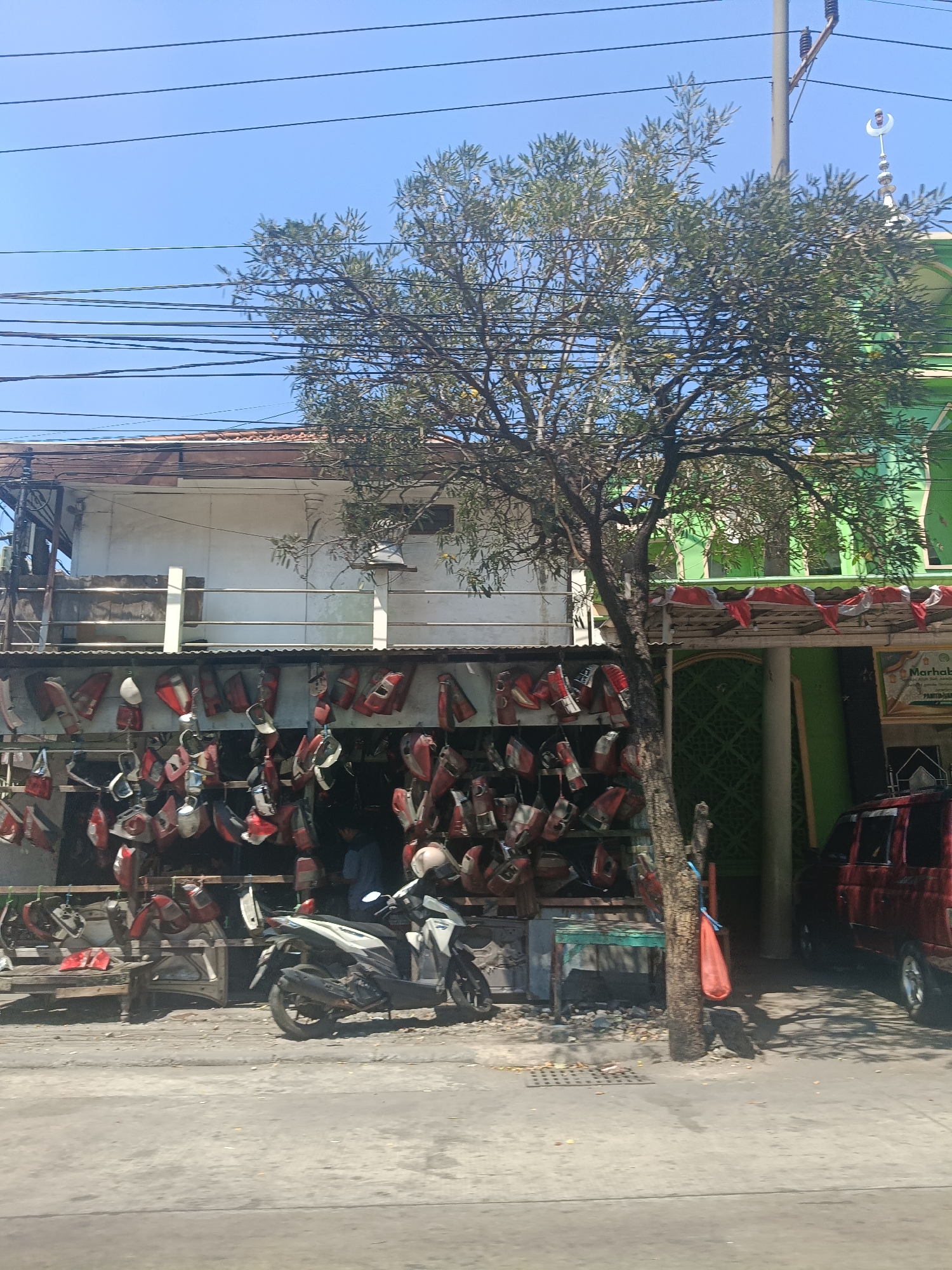 The image shows a street scene in Surabaya, Indonesia. A store is visible with motorcycle parts, specifically many red and white motorcycle fairings and panels, hanging on display. A white motorcycle is parked in front of the store. A red car is parked to the right of the store. A tree with sparse leaves grows in front of the store, and a green building with a crescent moon on top, resembling a mosque, is visible behind the tree and store. The sky is clear and blue. Power lines crisscross the top of the image. The scene appears to be a commercial area with a mix of residential and business establishments.