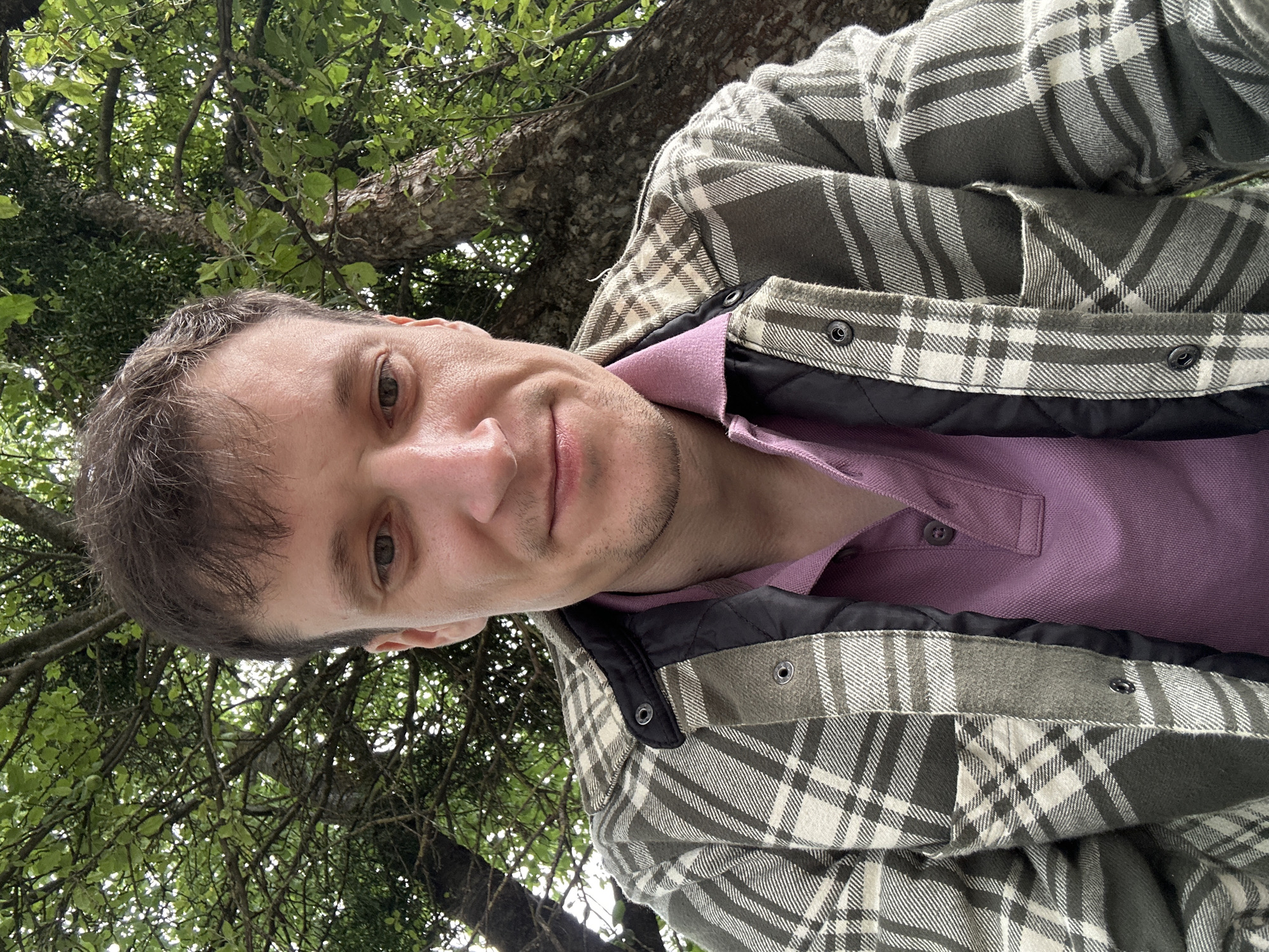 A man is sitting outdoors under a tree. He is wearing a plaid shirt over a purple polo shirt. The background consists of the tree's trunk and green foliage. The man's facial expression is neutral. The image appears to be a selfie, taken from a low angle. There is no discernible information about the city of Makiv, Ukraine in the image.