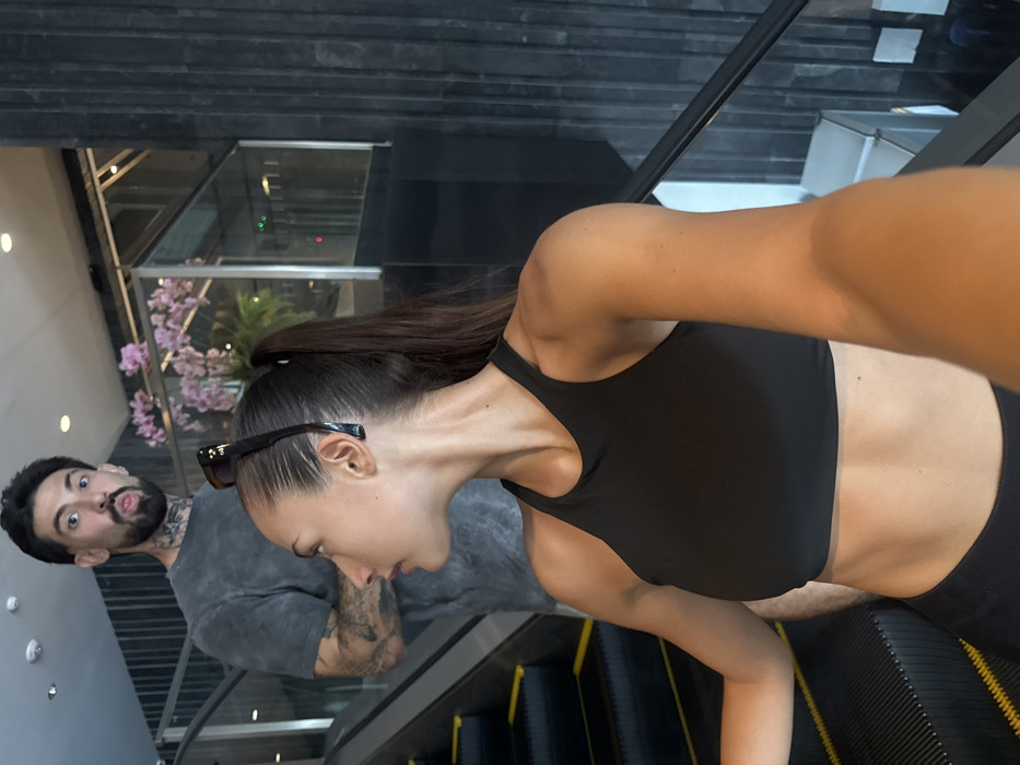 A female subject, seen in profile from the waist up, occupies the foreground, extending her right arm to capture a selfie. She has dark hair styled in a high ponytail, with dark-framed sunglasses perched on her head, and wears a black sports bra. Her gaze is directed slightly to her left. Immediately behind her and ascending on the same escalator, a male subject is visible from the chest up. He has dark hair, a beard, neck tattoos, and is wearing a grey t-shirt. His mouth is open, and his tongue is slightly extended, indicating a playful or surprised expression directed toward the camera. Both individuals are on an upward-moving escalator with black steps and yellow safety lines. The environment features a modern interior with dark, vertically paneled walls, glass railings, and a floral display of pink orchids behind a glass partition in the background. Recessed ceiling lights are also visible. This scene takes place within a modern commercial or public building in Bangkok, Thailand, likely a shopping mall or hotel, though the precise location is not definitively identifiable from the image.