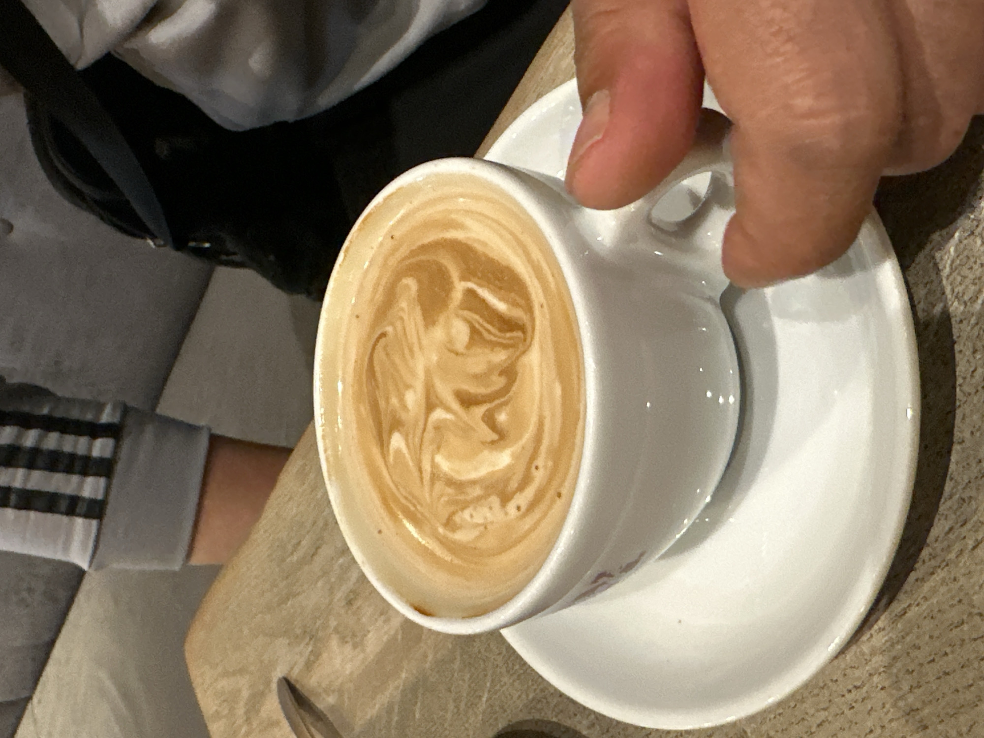 A close-up shot shows a hand holding a white cup of coffee with latte art on a saucer. The coffee appears to be a cappuccino or latte, with a creamy brown foam artfully swirled on top. The cup and saucer are placed on a wooden table. In the background, a person wearing a grey shirt with black stripes and a black backpack can be seen sitting on a grey couch. The overall lighting suggests an indoor setting, possibly a cafe. The image does not provide any information that allows for the identification of a specific location in Krefeld, Germany.