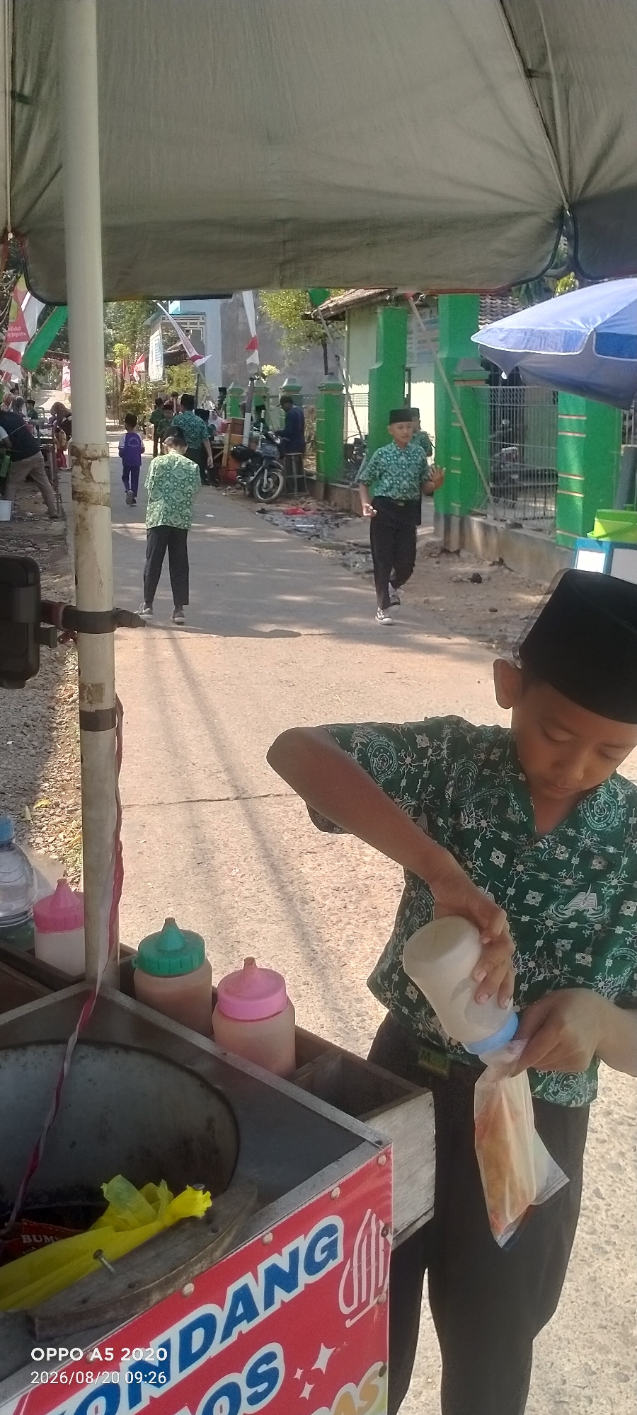 The image shows a young boy in a green patterned shirt and black pants, wearing a black peci (cap), pouring a white liquid from a bottle into a plastic bag. He is standing next to a food cart. The cart has a metal basin and several colorful bottles with lids on top. The side of the cart is adorned with red panels featuring white and colorful text and graphics. The scene appears to be outdoors, with a paved road visible.
In the background, other people, mostly children in similar green shirts and black pants, are walking or standing. Some are carrying school bags. There are also a few motorcycles parked and a building with green pillars and white walls, possibly a mosque or school. Indonesian flags are visible, suggesting a national holiday or event. The lighting suggests it is daytime, likely morning or afternoon, with bright sunlight casting shadows. The overall atmosphere seems lively and casual.
Visible text on the food cart includes "DANG" and "OS". At the bottom left of the image, there is a timestamp and device information: "OPPO A5 2020 2026/08/20 09:26". The location context is Sowan Kidul, Indonesia.