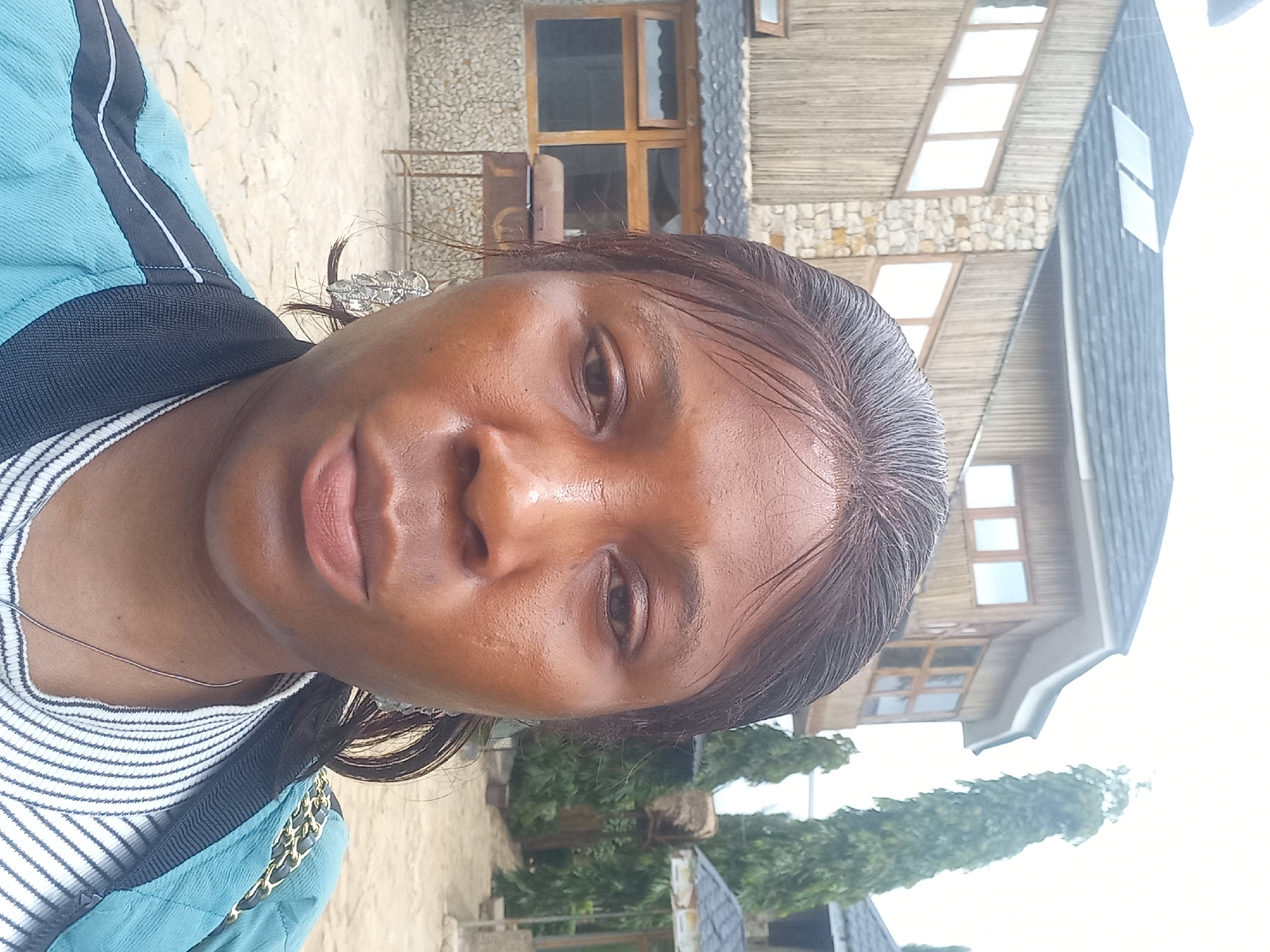 A medium-shot, eye-level, outdoor selfie features a woman in the foreground, facing the camera. She has dark skin, dark hair pulled back, and is wearing silver earrings. Her face is centered and takes up a significant portion of the frame. She is wearing a light blue bomber jacket over a striped shirt. The background reveals a building with a dark, slanted roof and walls constructed from bamboo and stone. Several tall, slender trees are visible to the right of the building, along with a cloudy sky. The ground is covered in light-colored gravel. The overall impression is that of a residential or resort setting.
This scene is located in Jos, Nigeria.