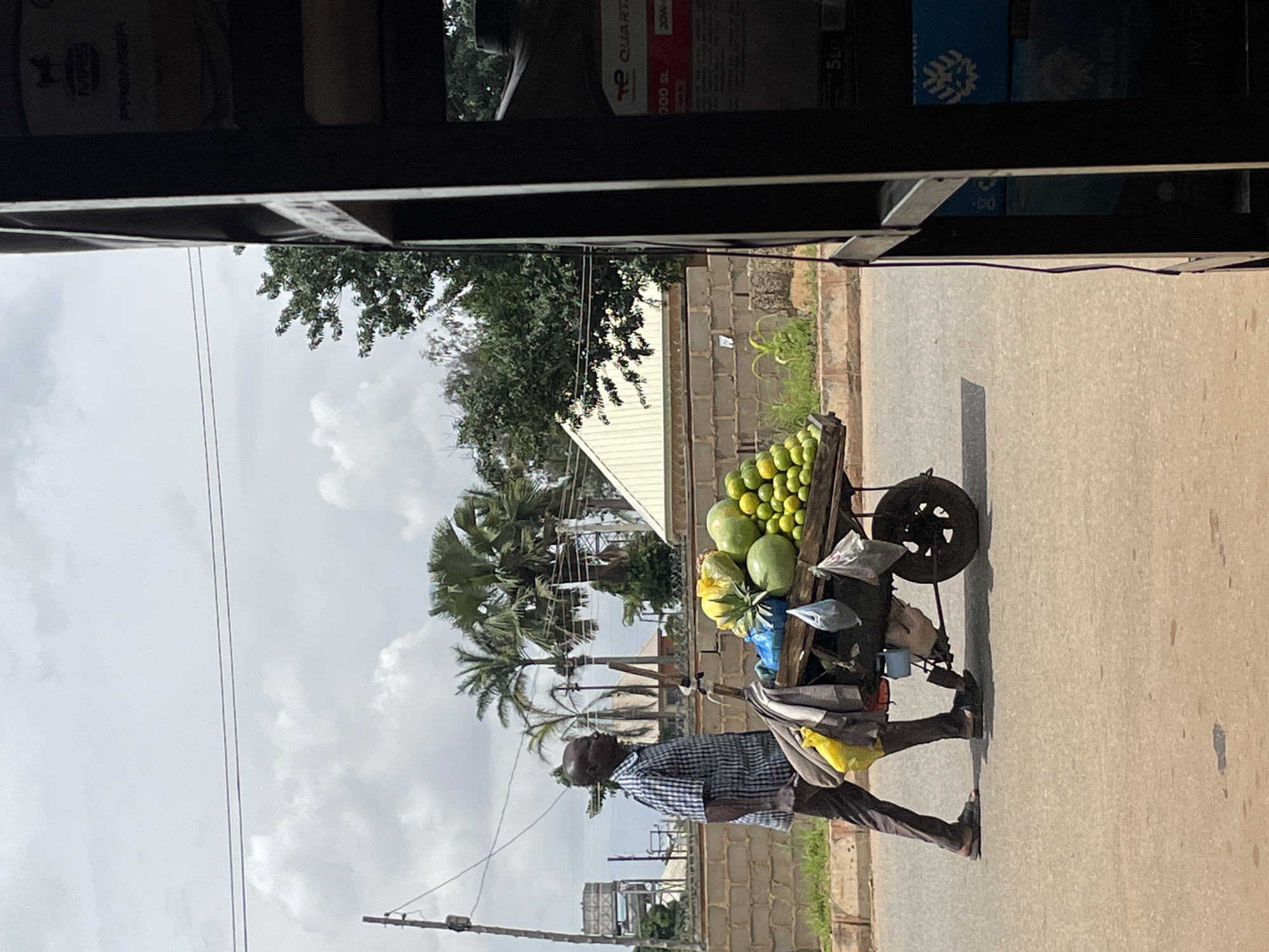 A man walks on a paved road in Bukuru, Nigeria, pushing a two-wheeled cart laden with fruit. The man, wearing a checkered blue and white short-sleeved shirt and dark brown trousers, has dark skin and is bald. He wears dark sandals. The cart is made of wood and metal and carries a variety of fruits including watermelons, oranges, and pineapples, along with some yellow and blue plastic bags and other items. The cart has a single wheel at the back. Behind the man, a brick wall runs along the side of the road, with vegetation and palm trees visible beyond. The sky is overcast with scattered clouds. On the right side of the image, a dark, metal shelving unit is partially visible, containing stacked boxes and containers, some with labels.