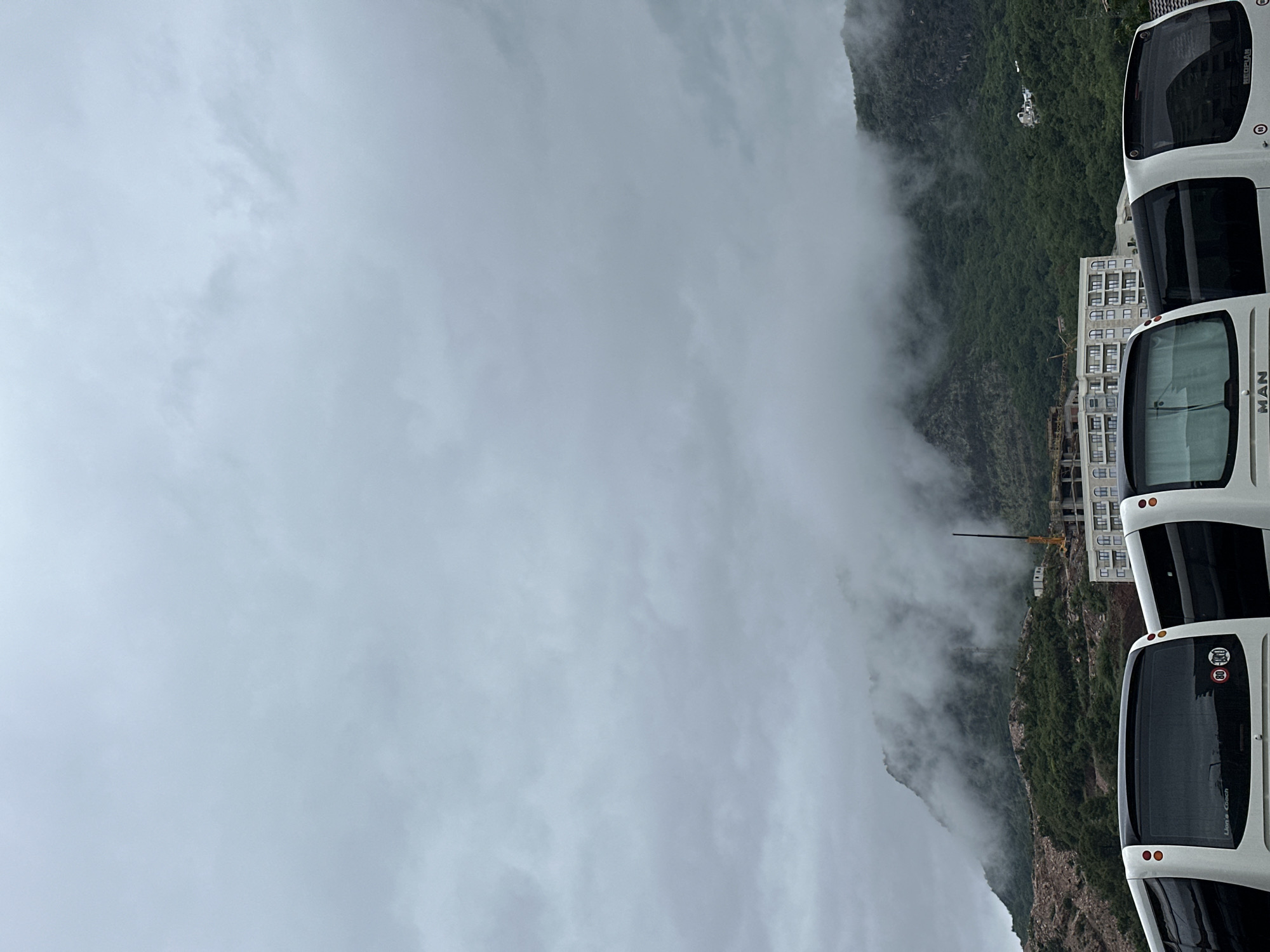 The image presents a foggy mountain landscape in Donji Pobori, Montenegro. The right side of the image shows the rear of three buses, one marked with "MAN" and another with "Lion's Coach." Between the buses and the mountain are a construction site with a yellow crane and multi-story buildings. The mountain is partially obscured by fog. The sky is uniformly overcast.