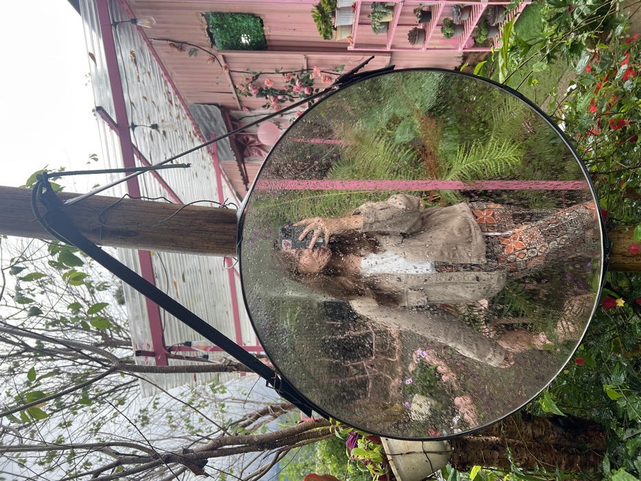 The image presents an outdoor scene located in Đưng K'Si, Vietnam. A large, circular mirror, approximately 0.8 meters in diameter, with a black frame and covered in water droplets, is centrally positioned. It hangs from a stout wooden pole via a black strap. The mirror reflects a female subject, observed from the waist up. She possesses long, wavy brown hair, wears a light brown or beige button-up jacket over a white undershirt, and patterned pants featuring orange and dark motifs. The subject holds a black smartphone to her face, capturing her reflection, with a metallic ring visible on her right hand's ring finger.
The surrounding environment consists of lush, dense greenery. To the left, a tree with thin, leafless branches extends upwards against an overcast, grey sky. A white hanging pot with green foliage is attached to a smaller tree trunk. Behind the mirror, various green plants, including ferns, are visible. To the right and in the background, a distinctive structure constructed of pink corrugated metal is present. This building features a pitched roof with exposed pink support beams, and is decorated with string lights containing Edison-style bulbs. A large pink bow and artificial pink roses adorn its facade. A square opening in the wall is filled with green artificial foliage. Adjacent to the building on the right, a pink multi-tiered shelving unit displays an array of potted plants, including succulents and small bonsai trees. Red and green flowering plants are observed in the immediate foreground, contributing to the vibrant garden setting.