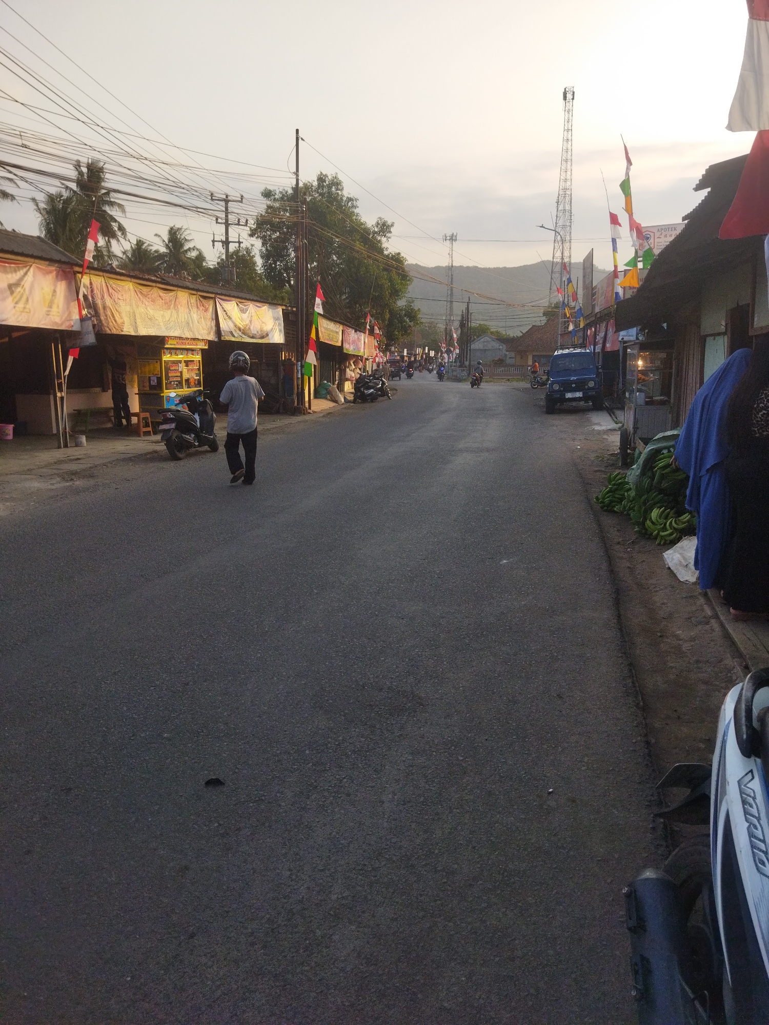 The image shows a street scene in Banjar, Indonesia, likely during daylight hours. The street is paved with asphalt and has a slight incline. On both sides of the street are small shops with awnings. Numerous Indonesian flags are displayed, suggesting a patriotic or festive occasion.
In the foreground, a person wearing a light-colored shirt and dark pants walks away from the camera. A motorcycle is parked to their left. Further down the street, several other motorcycles are visible, some being ridden. A blue utility vehicle is parked on the right side of the road. A large pile of bananas is situated next to a shop on the right.
In the background, there are trees, power lines, and a distant view of hills. Two cell towers are prominent in the skyline. The sky is overcast, indicating a possibly cloudy or hazy day. The overall impression is of a bustling local street in a semi-urban or rural area.