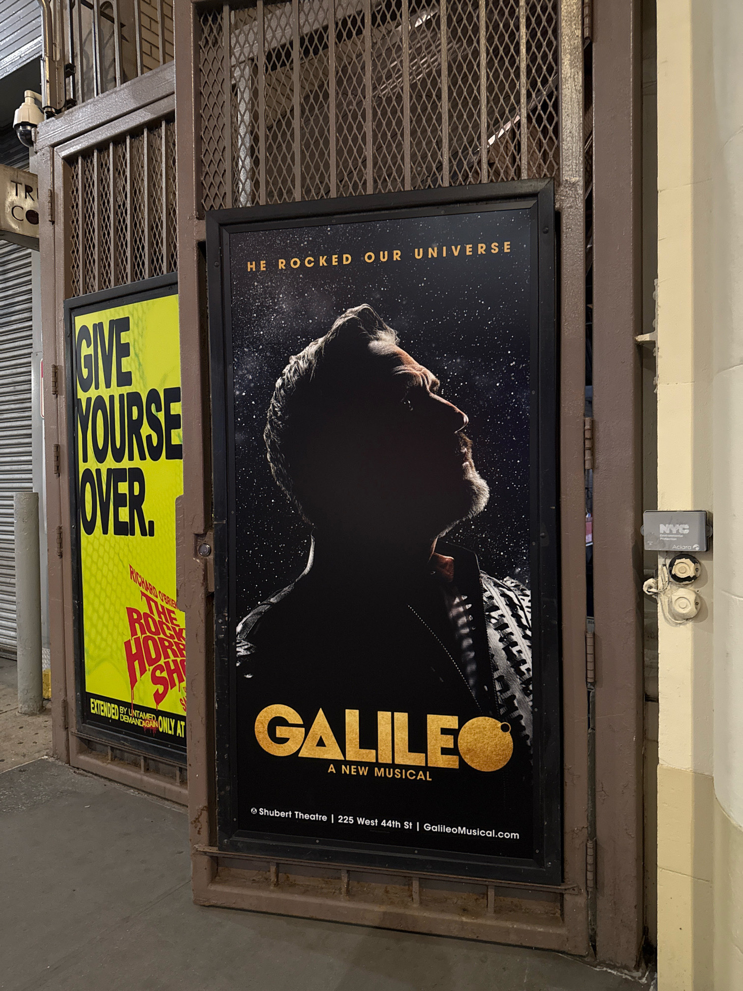 The image shows an exterior view of a building in New York City, United States of America, likely a theater. Two posters are prominently displayed on a metal gate. The poster on the right advertises a musical titled "GALILEO: A NEW MUSICAL" with the tagline "HE ROCKED OUR UNIVERSE." The poster features a silhouette of a man looking upwards against a starry background. Below the title, it states "Shubert Theatre | 225 West 44th St | GalileoMusical.com." The poster on the left is bright yellow with black text that reads "GIVE YOURSELF OVER." Below this, smaller red text mentions "RICHARD O'BRIEN'S THE ROCKY HORROR SHOW" and "EXTENDED BY UNTAMED DEMAND AGAIN ONLY AT." The posters are attached to a dark brown metal gate with vertical bars and diamond-shaped mesh. A security camera is visible above the gate on the left. The building facade on the right is painted a light cream color, and a gray NYC Department of Transportation device is mounted on the wall. The ground in front of the gate appears to be a sidewalk.