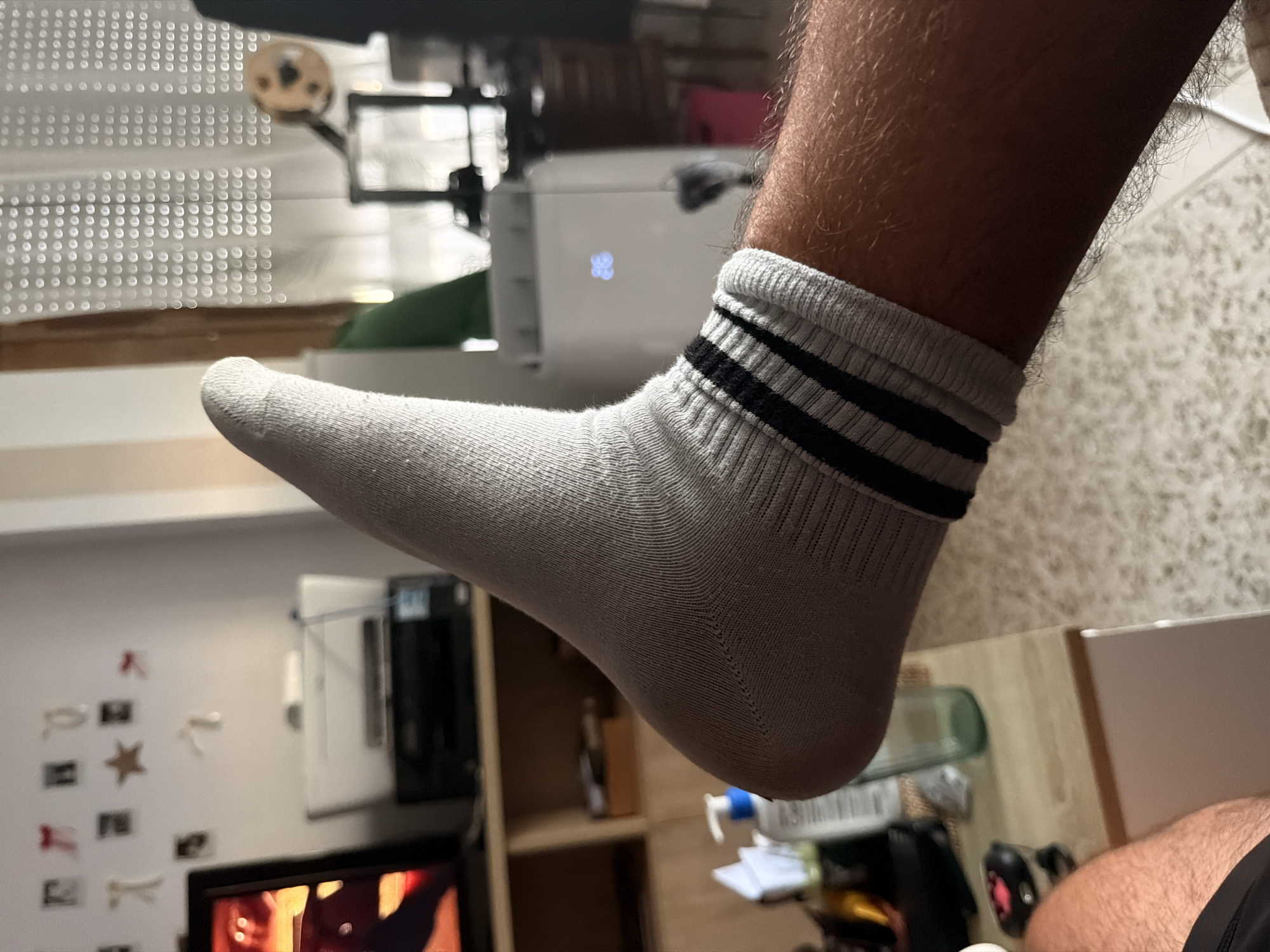 The image shows a close-up of a person's right foot wearing a grey sock with black stripes. The foot is extended upwards and to the left, with the heel facing the viewer. The sock appears to be made of a ribbed material, and the black stripes are positioned around the ankle area. The person's leg, covered in dark hair, is visible below the sock.
In the background, a wooden shelf unit is partially visible, holding a printer and what appears to be a television screen showing a brightly colored image. To the right, a white portable air conditioner unit is present, with a blue digital display showing a snowflake icon. Behind the air conditioner, sheer curtains with a circular pattern cover a window, and a wooden chair with a red cushion is visible. The environment suggests an indoor residential setting.
The image is focused on the foot and leg, with the background elements out of focus. The lighting appears to be artificial, casting shadows and highlighting the texture of the sock and the hair on the leg. There is no information in the image to indicate the location as Castillo Bajo-conejito, Spain.
