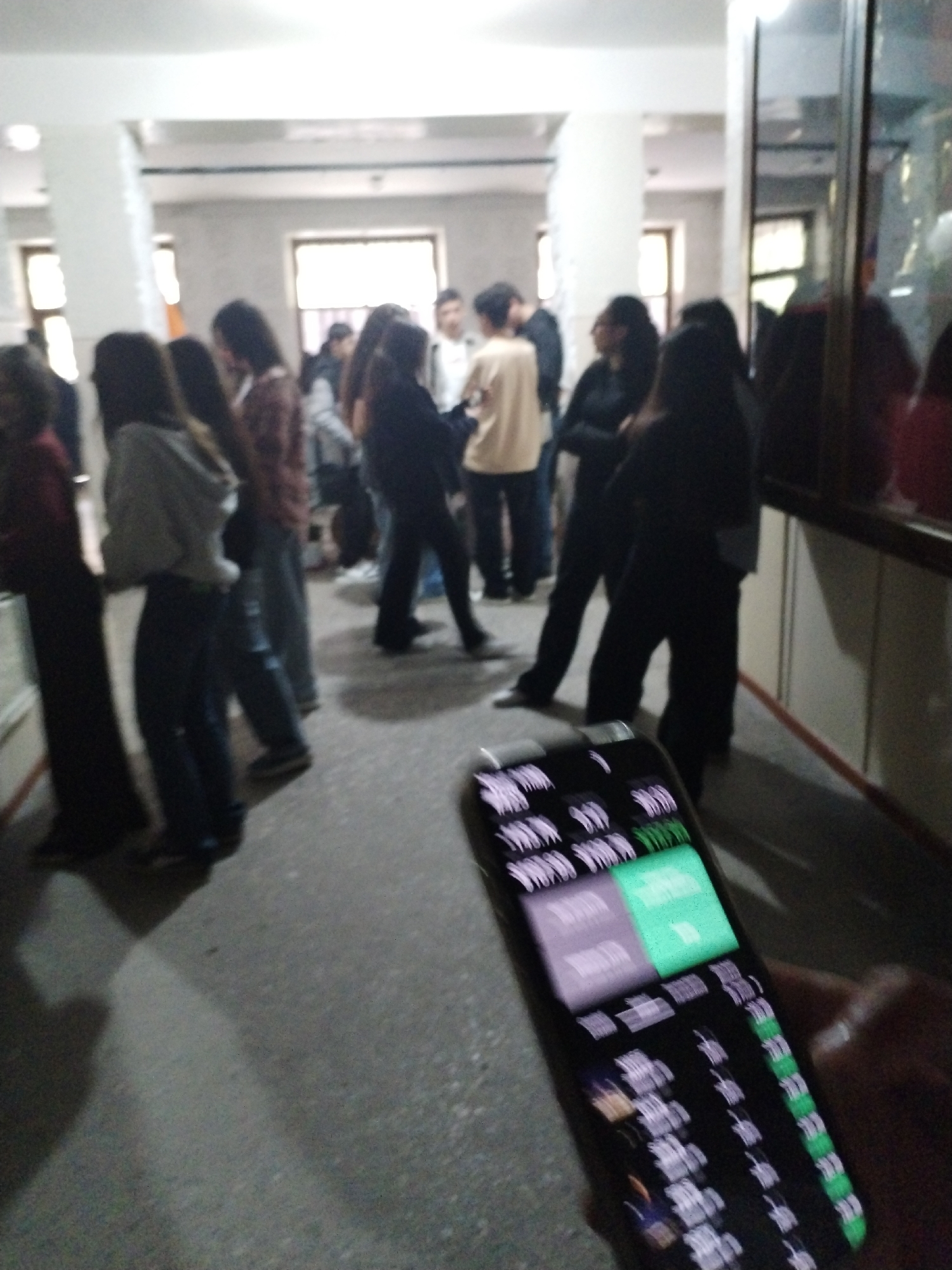 The image captures an indoor scene, likely a hallway or large room within an institutional building such as a school or university. A group of approximately ten to fifteen individuals, primarily young adults or teenagers, are gathered and standing in various small clusters. Their faces and expressions are largely indistinct due to the image's blurriness and distance.
In the immediate foreground, a hand holds a smartphone, which is angled towards the viewer. The phone's screen displays what appears to be a list-based interface with both text and numerical data. Several lines of text resembling "WE W W W W W" and "WWW" are visible, interspersed with what look like numerical values, some highlighted in green, possibly indicating active status, positive change, or available quantities. One prominent section of the interface is entirely green, suggesting a selectable option or a significant status.
The background features several large pillars and window-like structures, allowing natural light into the space, complemented by overhead artificial lighting. The flooring is a light-colored, possibly concrete or tiled, surface. The overall atmosphere appears casual, suggesting a waiting period, break, or social gathering. The time of day appears to be daytime, given the natural light. The primary activity depicted is casual standing and interacting among the group, while the person in the foreground is engrossed with their phone.