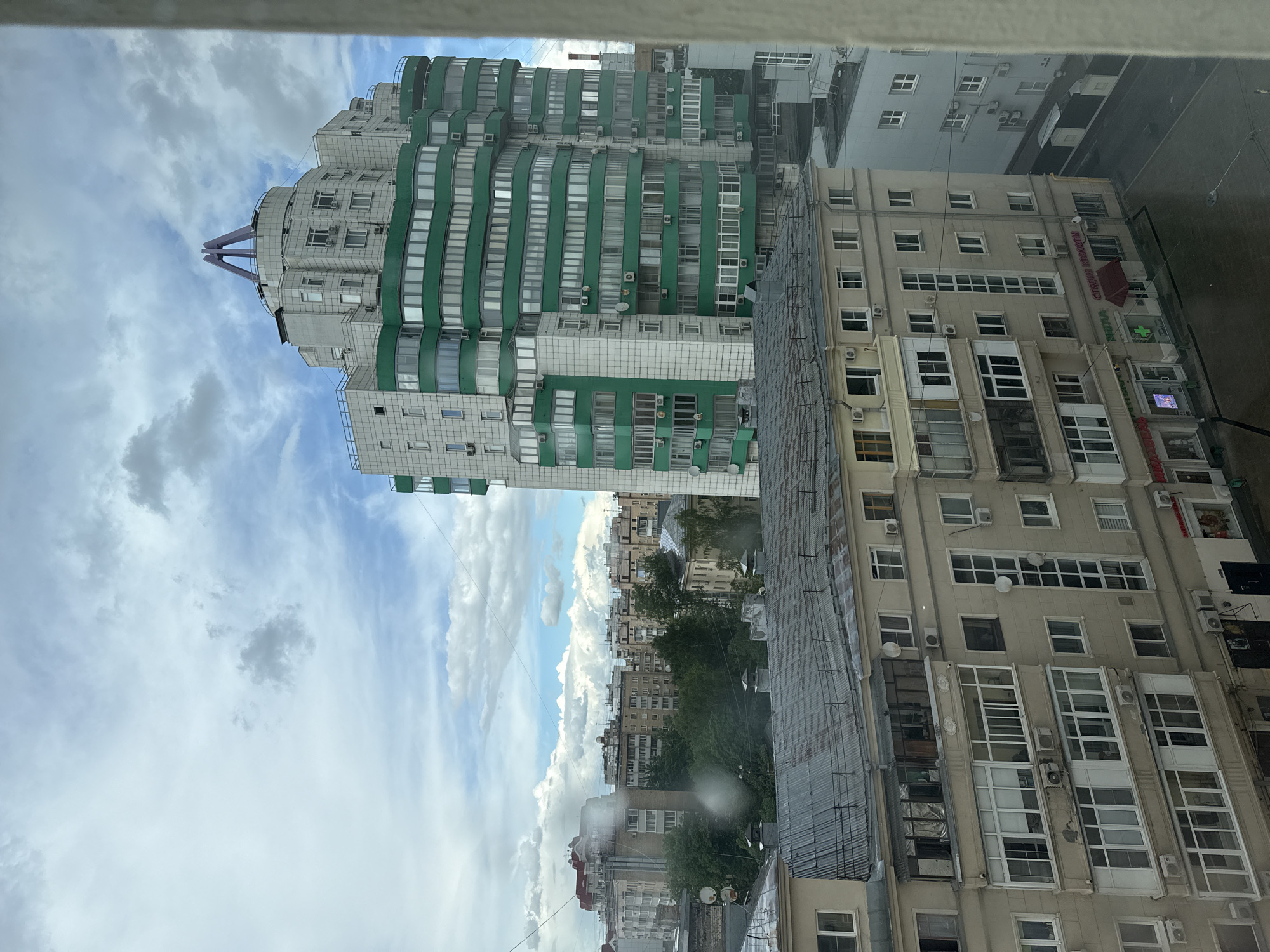 The image presents an elevated urban view, possibly from an apartment window. Prominent features include high-rise buildings: one with a striking green and white facade with distinct horizontal banding, and another older building is tan. Overhead, the sky is filled with billowing white clouds. The perspective suggests a location within the city of Moscow, Russia, as this architectural style is common in the area. Raindrops are visible on the window. There is no discernible human presence.