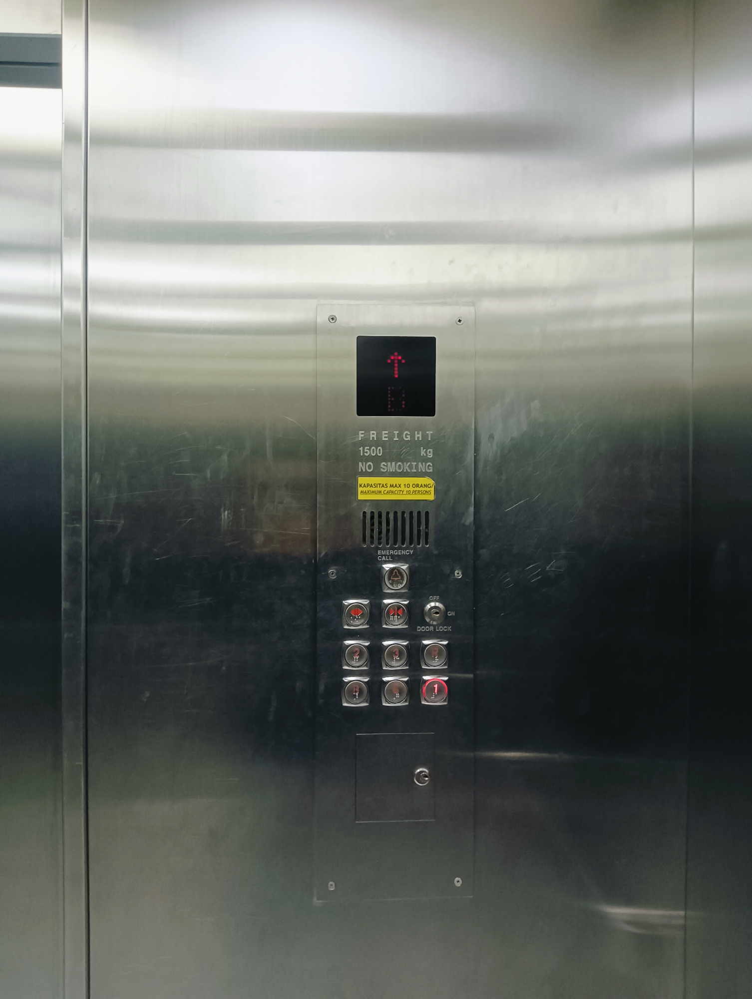 The image shows the interior of a brushed metal elevator. The elevator panel features a digital display showing an upward arrow and the letter "B" in red. Below the display, text indicates "FREIGHT 1500 kg NO SMOKING" and "KAPASITAS MAX 10 ORANG / MAXIMUM CAPACITY 10 PERSONS". Further down are several buttons, including "EMERGENCY CALL", "DOOR LOCK" with an on/off switch, and numbered buttons for floors. The number "1" button is illuminated in red. The overall environment appears to be a service or freight elevator. The context suggests this is likely a location in Batam, Indonesia, given the Indonesian text on the capacity warning.