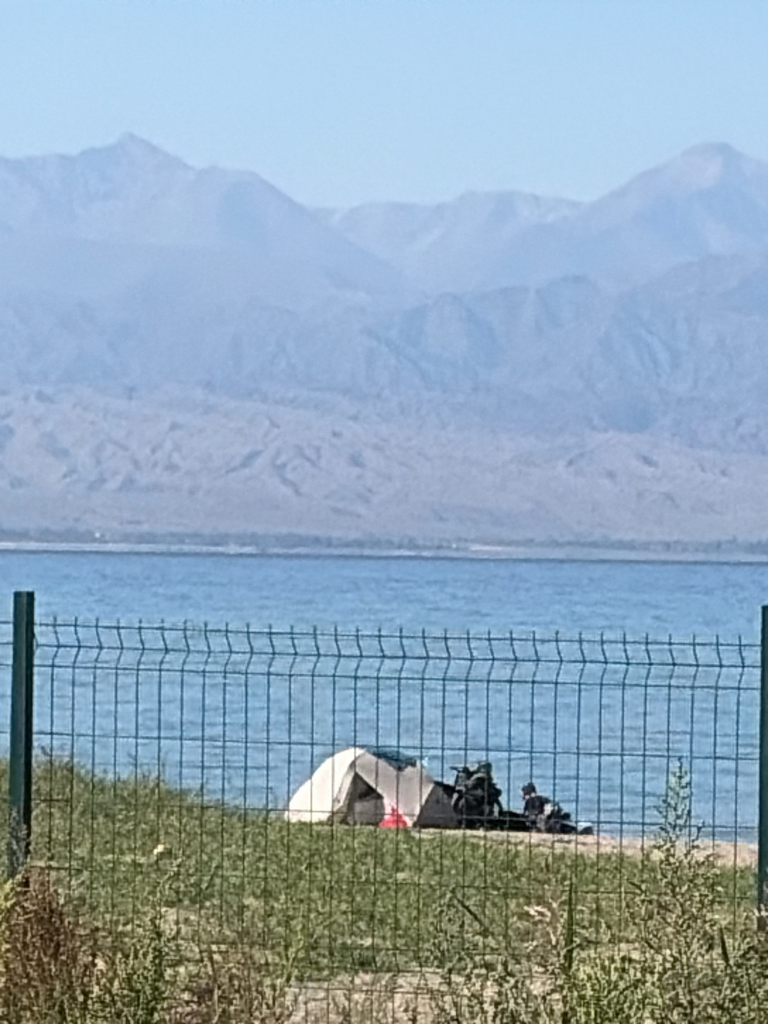The image shows a body of water with mountains in the background. In the foreground, there is a fence and a grassy area with a tent set up. Two people are seen near the tent, one of whom is sitting. The scene appears to be a campsite by a lake or sea, with a scenic mountain backdrop.
