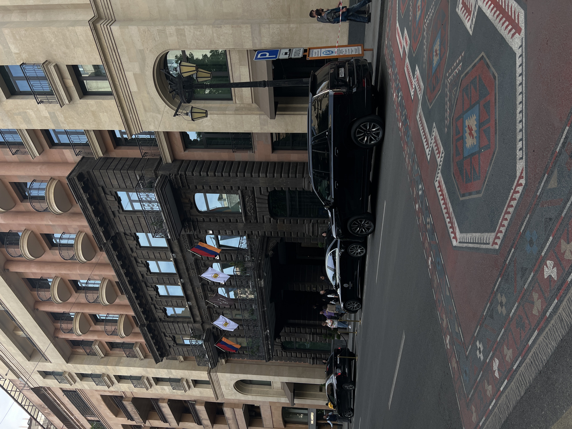 The image captures a street scene in Yerevan, Armenia, showcasing the exterior of The Alexander, a Luxury Collection Hotel. The hotel's facade is adorned with flags of Armenia and the European Union. Several black cars are parked along the street. The road features a painted carpet design. People are visible near the hotel entrance. Adjacent buildings with balconies and architectural details are also present.