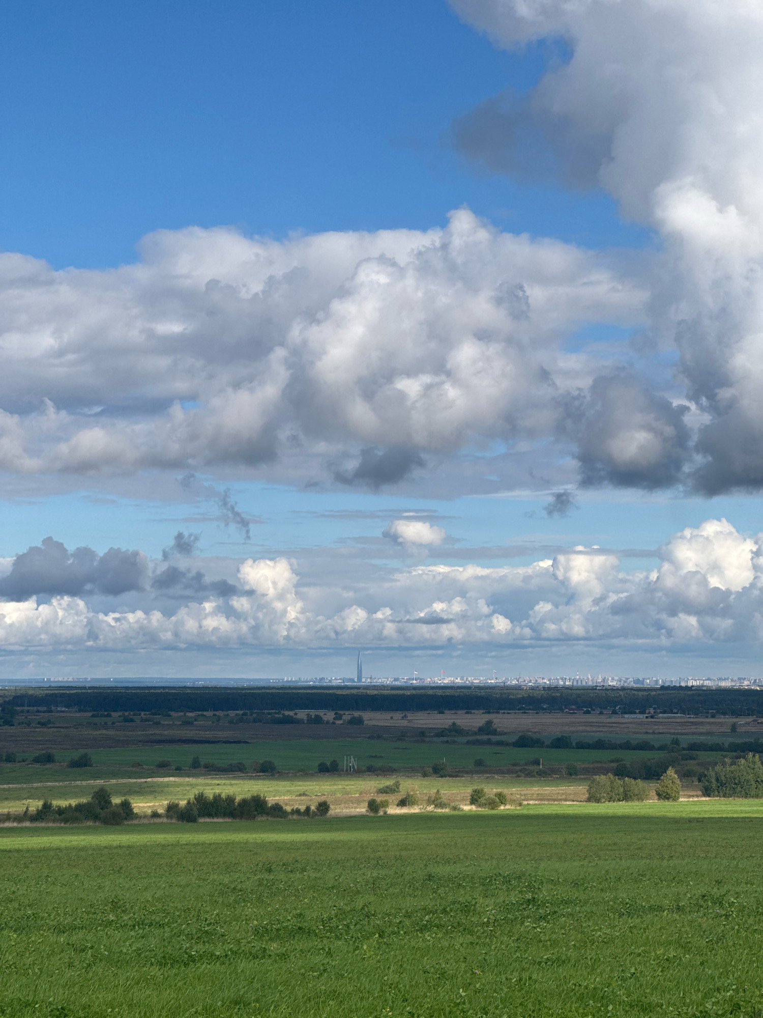 The image shows a panoramic view of the city of Kaporskoe, Russia, on a cloudy day. The foreground is dominated by a vast, lush green field, with gentle slopes and shadows indicating sunlight filtering through the clouds. In the mid-ground, there is a strip of dark green forest, followed by a patchwork of agricultural fields and scattered trees. The horizon is marked by the distant cityscape of Kaporskoe, characterized by numerous buildings and a prominent, tall skyscraper. The sky is a dramatic mix of bright blue patches and large, fluffy white and grey clouds.