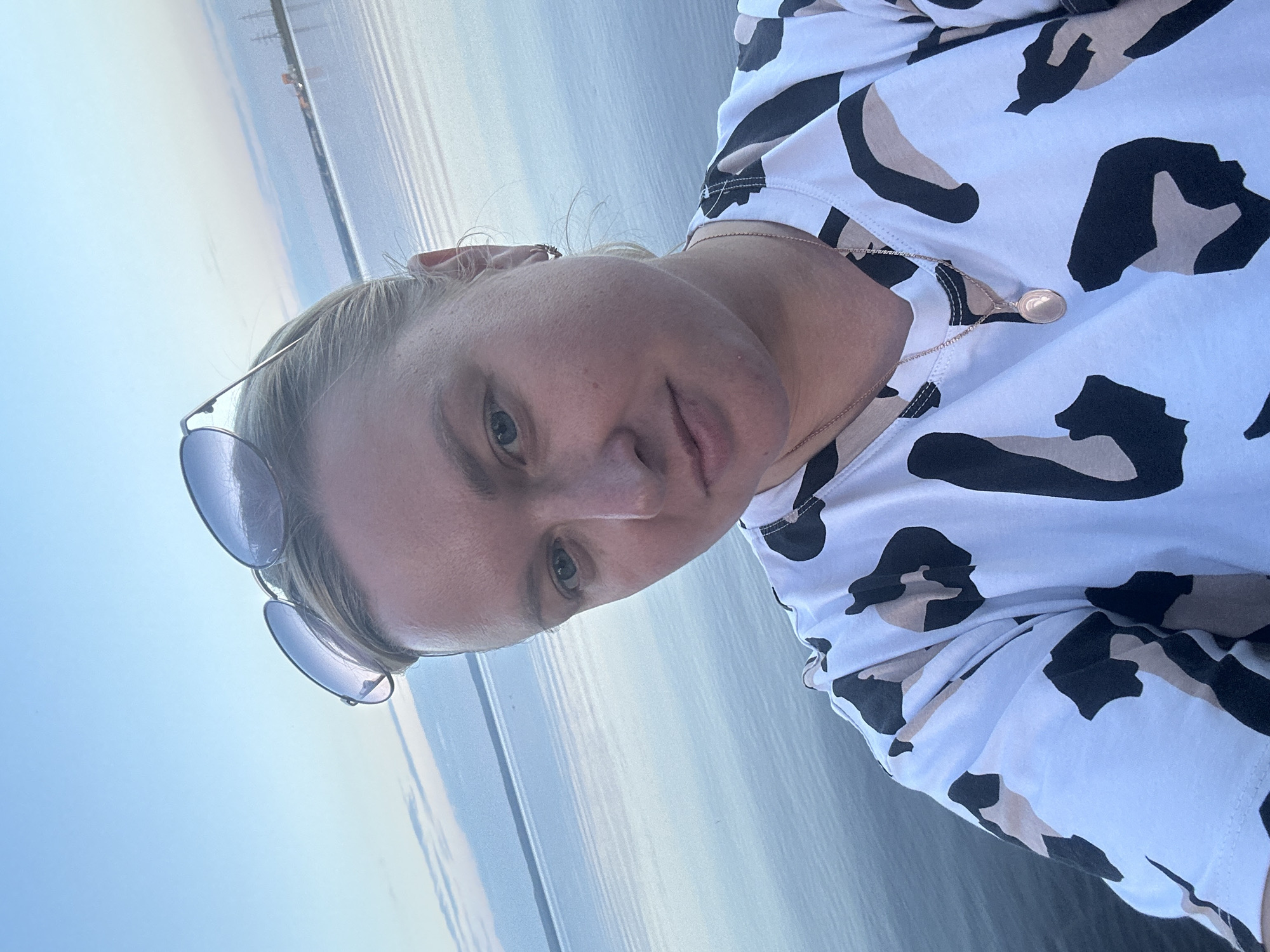 A selfie captures a blonde woman with sunglasses perched on her head, looking towards the camera. She is wearing a white t-shirt with a black and tan leopard print pattern and a delicate necklace with an oval pendant. The background reveals a wide expanse of calm water under a pale blue sky, likely a body of water in or near the city of Noviy, Russia. The water's surface reflects the sky, creating a serene and expansive atmosphere. In the distance, the faint outlines of industrial structures and power lines are visible on the horizon. The lighting suggests it may be late afternoon or early evening.