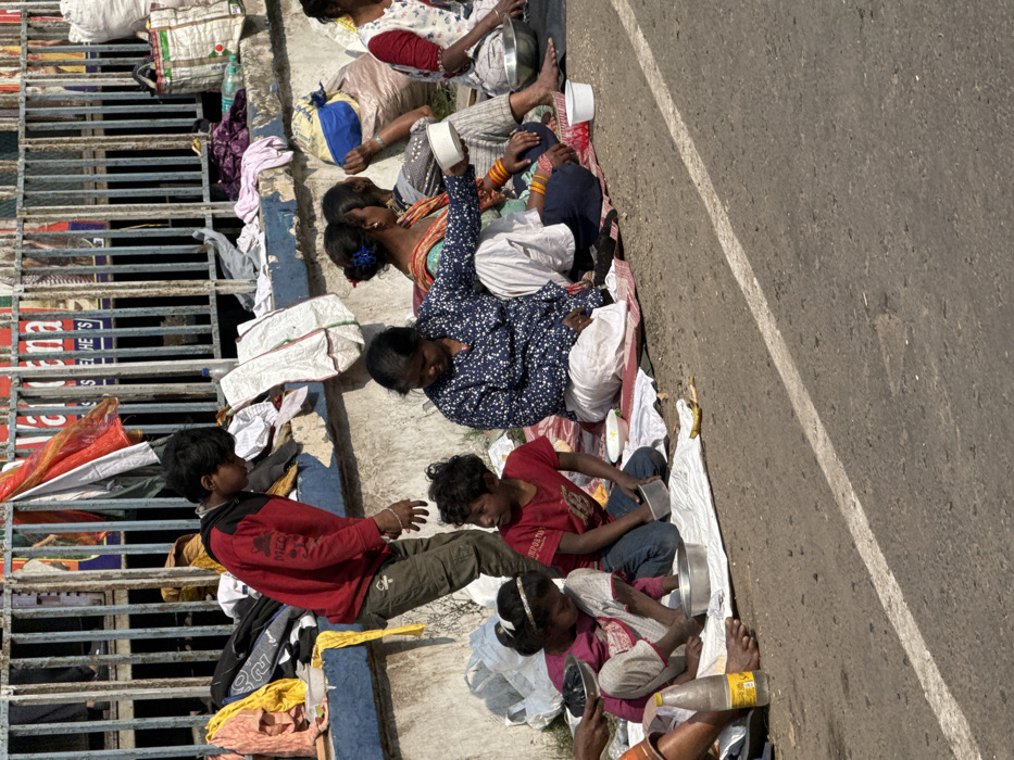 A group of people sit and lie alongside a road next to a guardrail. They appear to be impoverished, surrounded by bags, blankets, and personal belongings. Several children are playing and interacting with each other. Some people hold bowls, potentially seeking donations. The group sits directly adjacent to a grey asphalt road, indicating a precarious living situation.