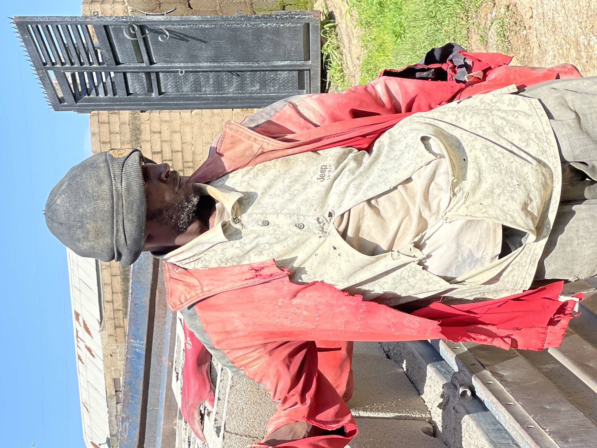 The image depicts a man in Jos, Nigeria, wearing a worn-out beanie and a red, tattered jacket over a distressed, patterned shirt. The man appears to be in an outdoor industrial or construction area, with a large metal gate and brick buildings in the background. Concrete blocks are visible in the foreground, suggesting construction materials. The overall scene conveys a sense of hardship and labor.