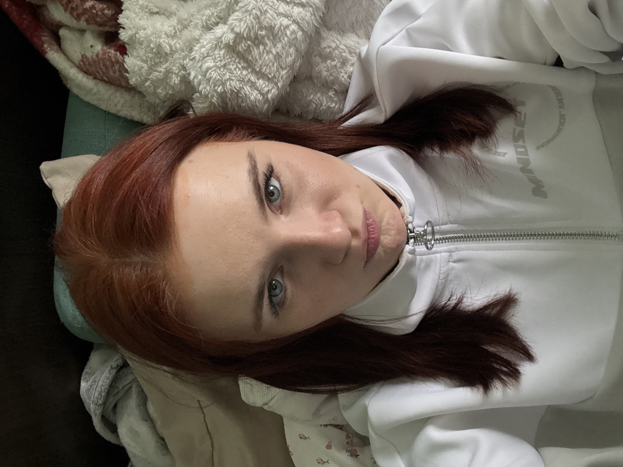 A woman with red hair and blue eyes is lying down wearing a white zip-up sweatshirt with "MNDSET" printed on it. She is surrounded by blankets and appears to be indoors. The city of Murino, Russia is not visible in the image and cannot be determined as the location.