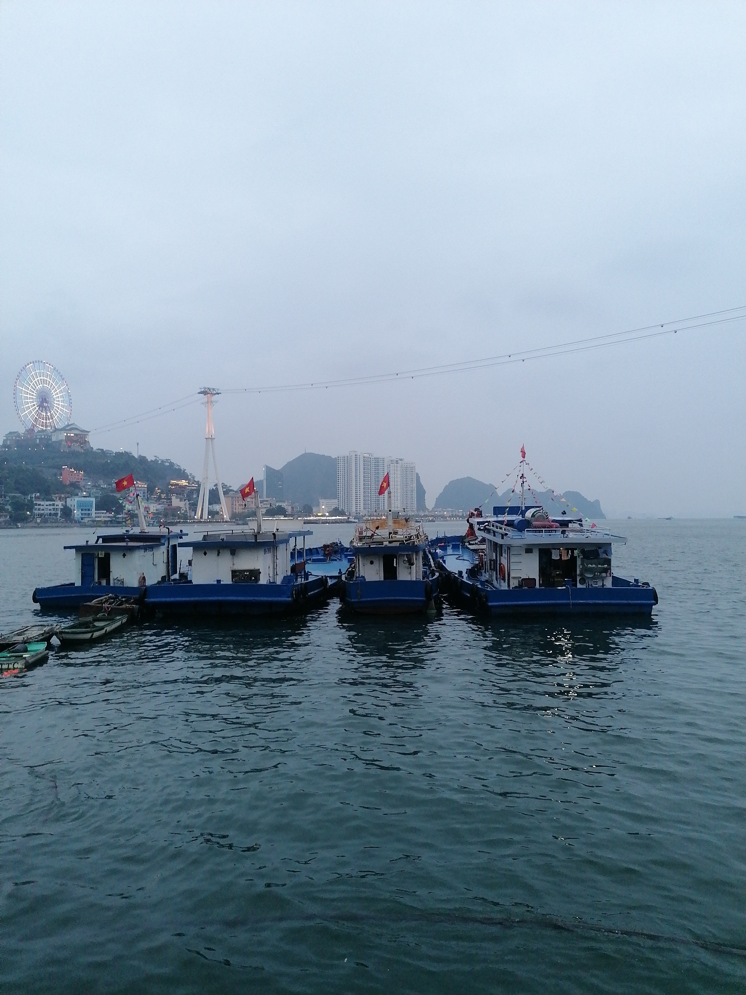 The image depicts a group of blue and white boats clustered together on the water near Bai Chay Ward, Vietnam. In the background, a Ferris wheel is visible on a hill, along with tall buildings and a tower connected by cables. Several Vietnamese flags are seen on the boats. Small rowboats are tied to the boats. The water is choppy with visible ripples. The sky is overcast.