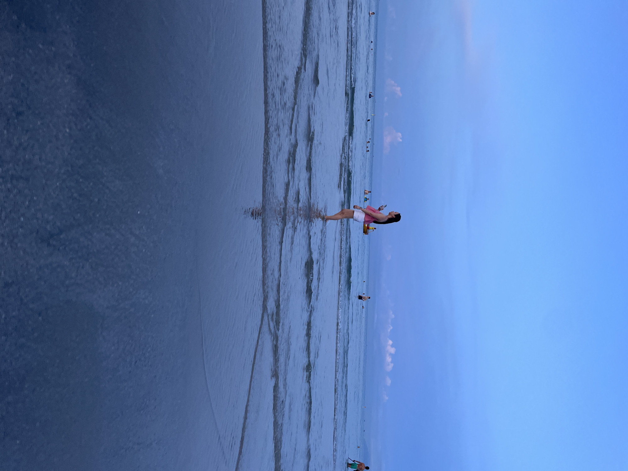 The image shows a beach scene with the ocean and sky. The orientation of the image is portrait mode, and the contents are rotated 90 degrees clockwise. A woman is standing in the shallow water near the shore, facing the ocean. She is wearing a pink top and white shorts and appears to be holding a phone or camera. Her body is angled, and she seems to be looking down. Several other people are visible in the distance, wading or swimming in the ocean. The water has gentle waves rolling in, creating rippled patterns on the wet sand. The sky is a clear, vibrant blue with a few wispy clouds. The lighting suggests it is either early morning or late afternoon, with a soft, diffused light. The overall impression is of a relaxed and peaceful day at the beach. The location is identified as Đà Nẵng, Vietnam.