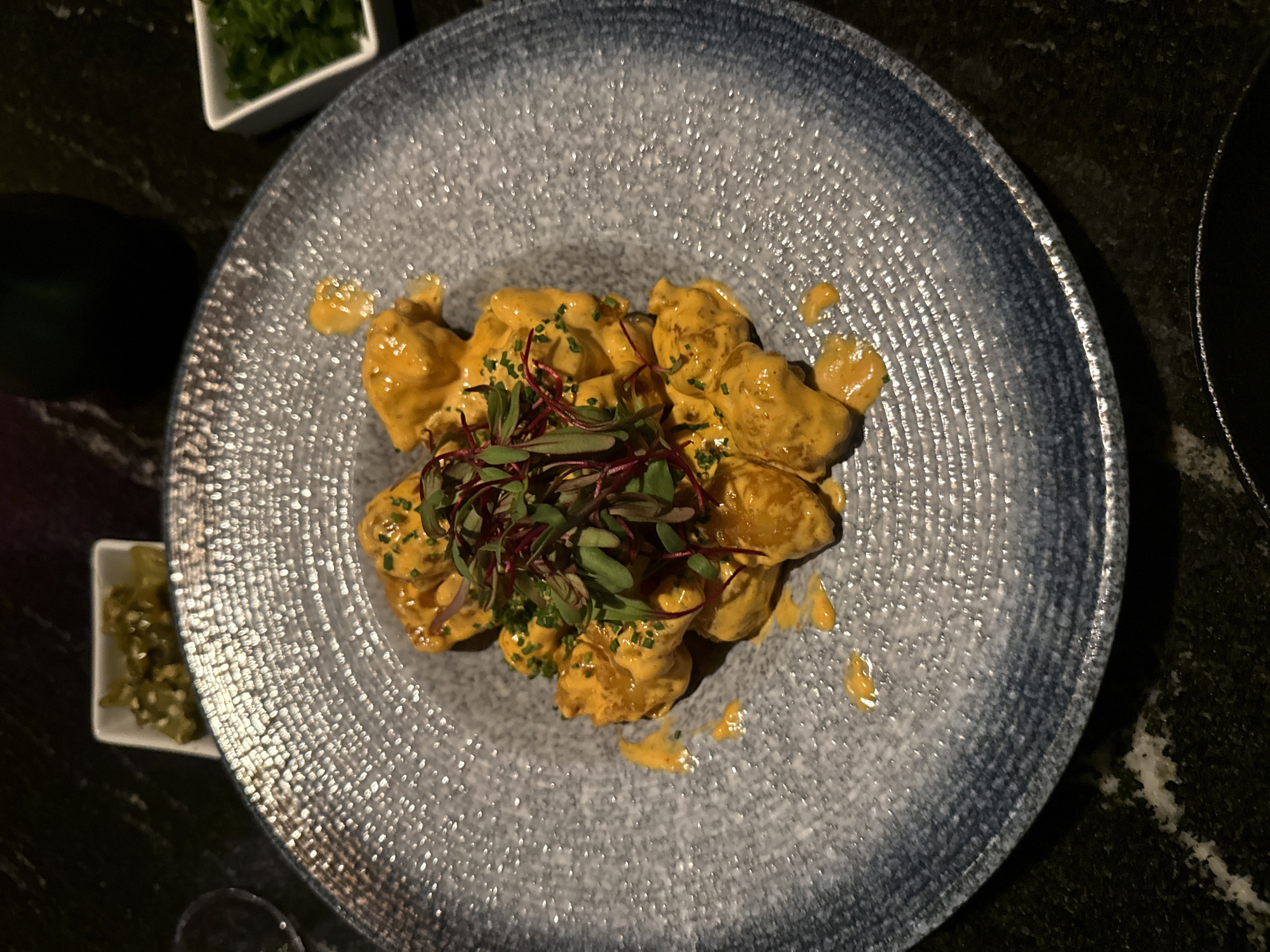 Overhead shot of a gray and blue textured plate with a mound of food covered in an orange sauce. The food is garnished with green and reddish-purple herbs. Small white dishes containing chopped green items are on the left. The plate is on a dark, possibly marble, surface. The location appears to be a restaurant setting, possibly in Mexico City, Mexico. No people are visible.