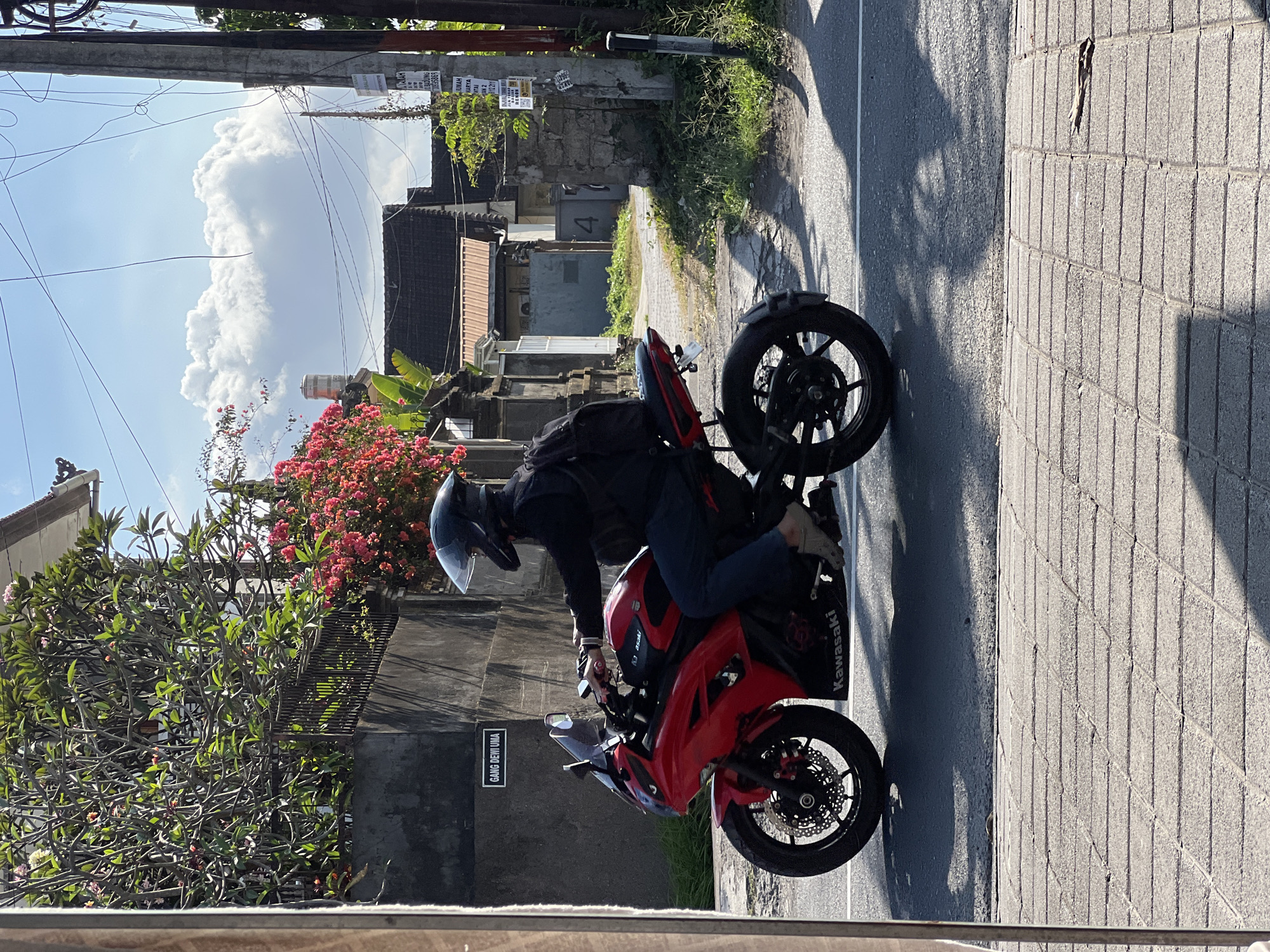 A person wearing a black helmet and black jacket is riding a red Kawasaki motorcycle on a street in Denpasar, Indonesia. The motorcycle is red with black accents. The rider is wearing blue jeans and light-colored shoes. Behind the rider, a black backpack is visible. The street has a checkered pavement on the side, and a road with a white line in the middle. In the background, there are buildings with tiled roofs, lush green trees, and bright pink flowers. A street sign with "GANG DEWI LIMA" is visible on a dark wall. The sky is blue with fluffy white clouds. Electrical wires are strung across the sky.