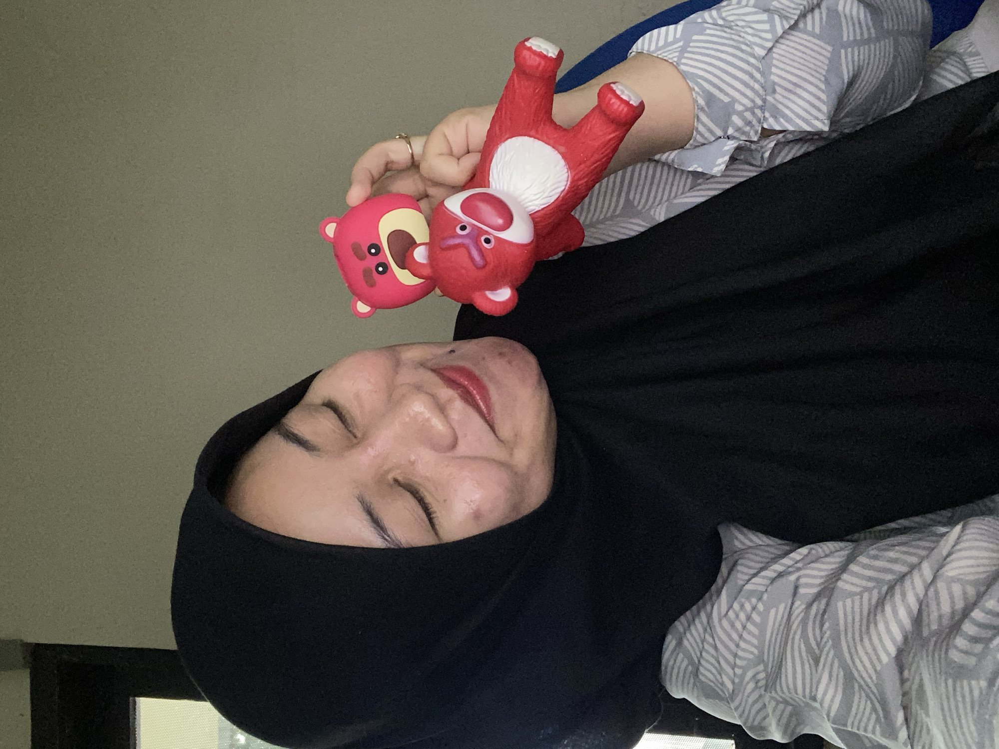 A woman wearing a black hijab and a gray and white patterned shirt is holding two Lotso Huggin' Bear toys. The woman's eyes are closed, and she is smiling. She is indoors, and the background is a plain, light-colored wall. The lighting is soft and natural. The image is a close-up, focusing on the woman and the toys. It is a selfie-style photograph. The location is unknown.
